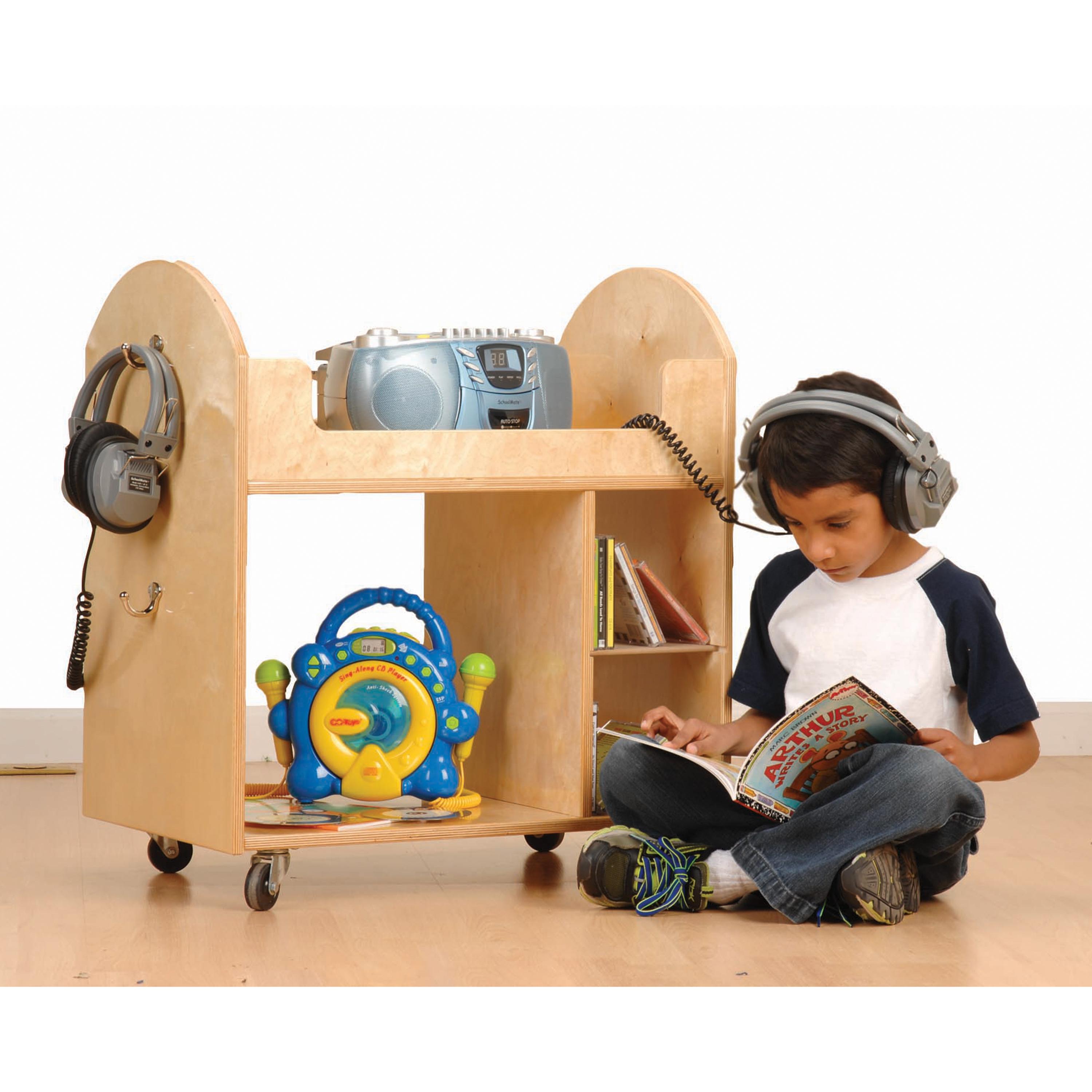 Child sitting on the floor with a book, wearing headphones, next to a wooden cart with toys and electronic devices.