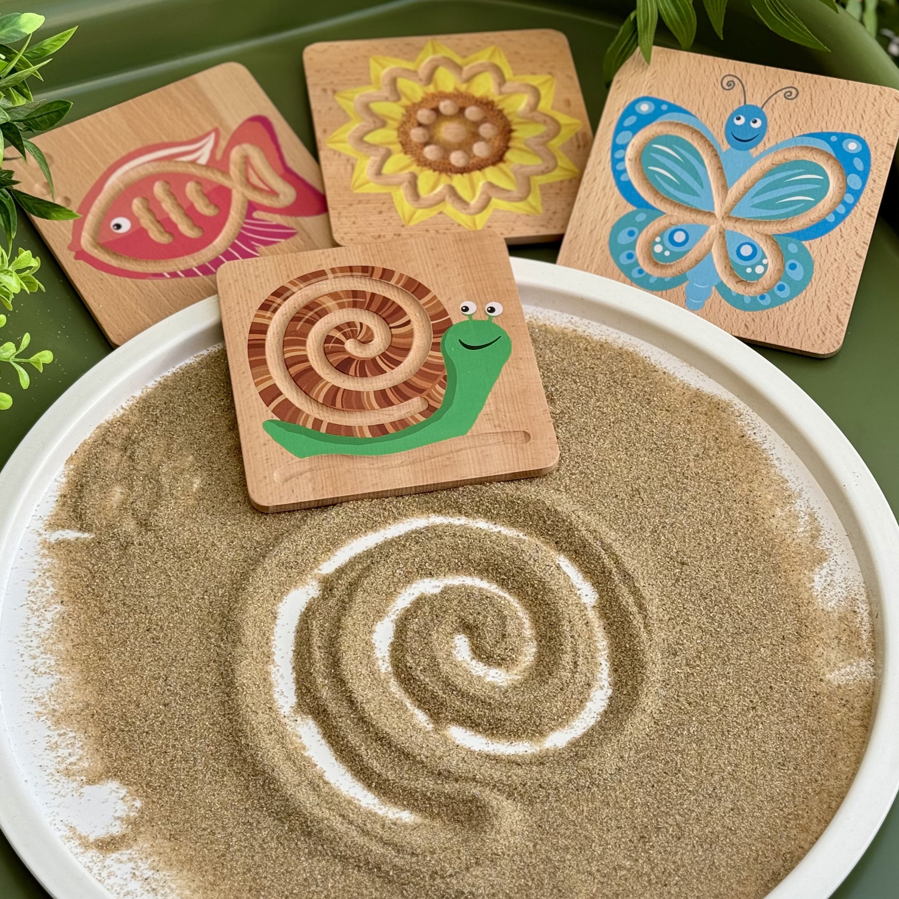 Wooden toys shaped like a fish, sunflower, snail, and butterfly on a sand tray with sand art.