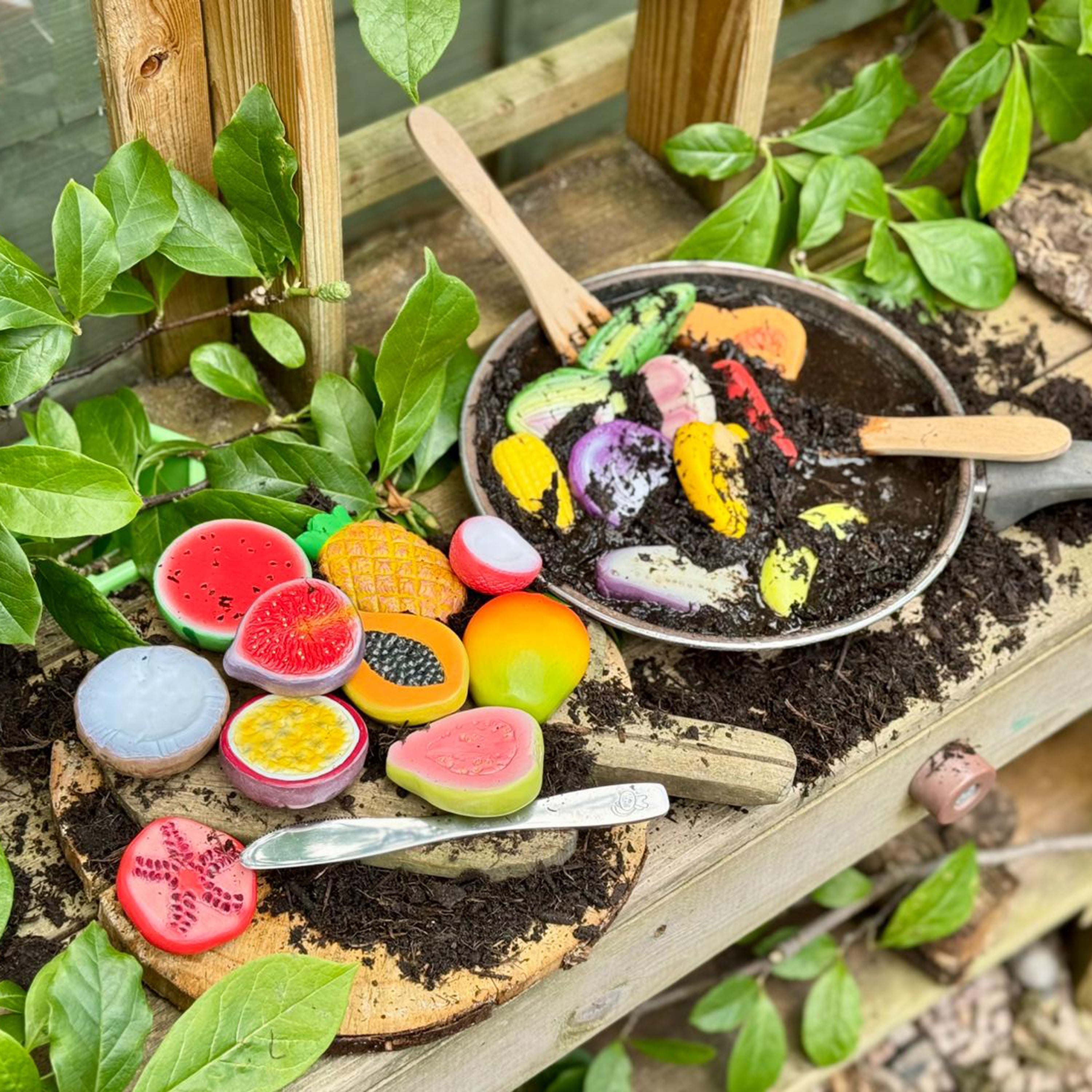Colorful toy fruits and vegetables in a pan with soil, surrounded by greenery.