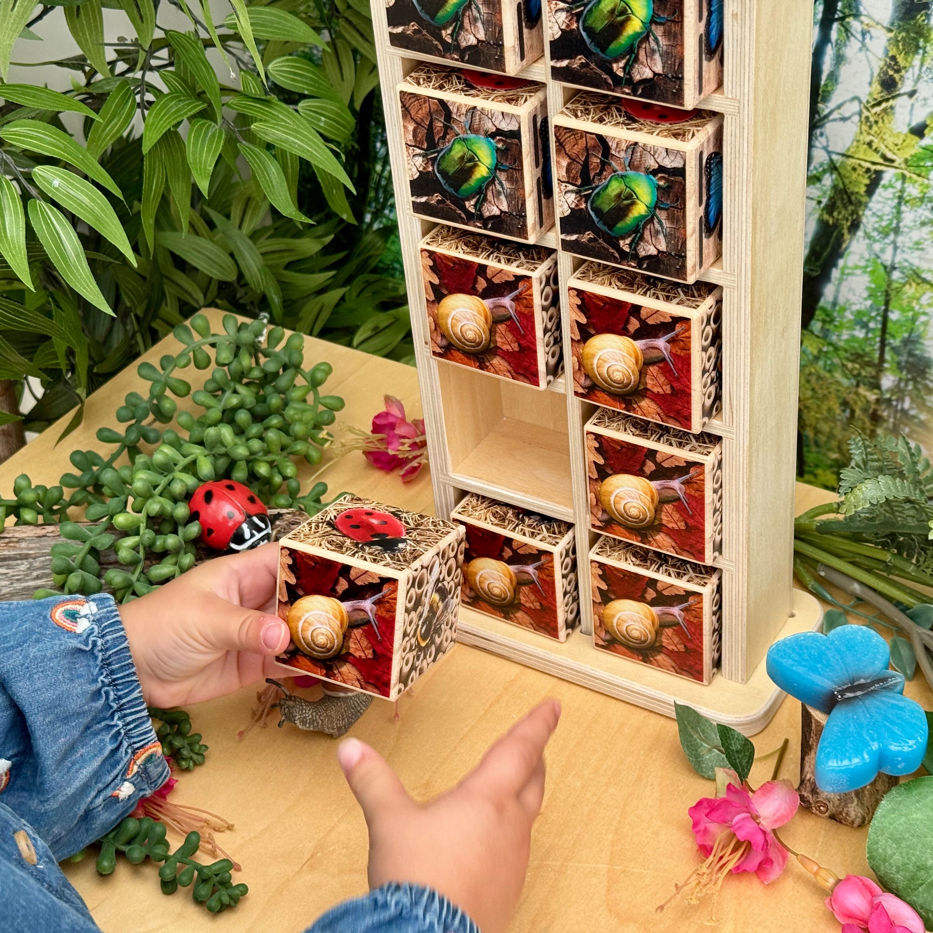 Wooden display case with small animals and plants on a wooden surface with greenery in the background.