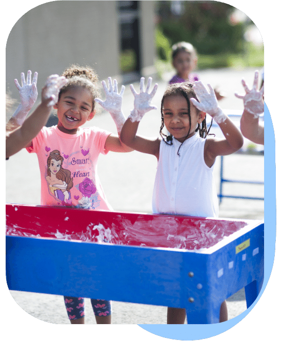 two girls hold their hands up covered in shaving foam as they play at the water table outside