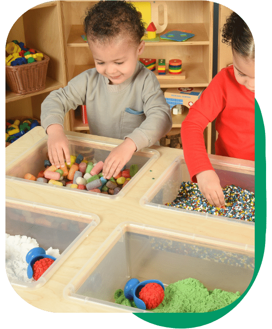 two children playing at the mobile sand and water table featuring four clear bins full of sensory materials- sand, pellets, rollers, etc.