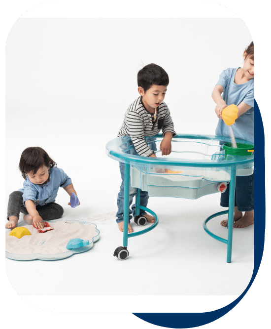 two children standing at the sand and water clam table playing with scoops and sand. another child sitting on the floor playing with sand in the removable tray