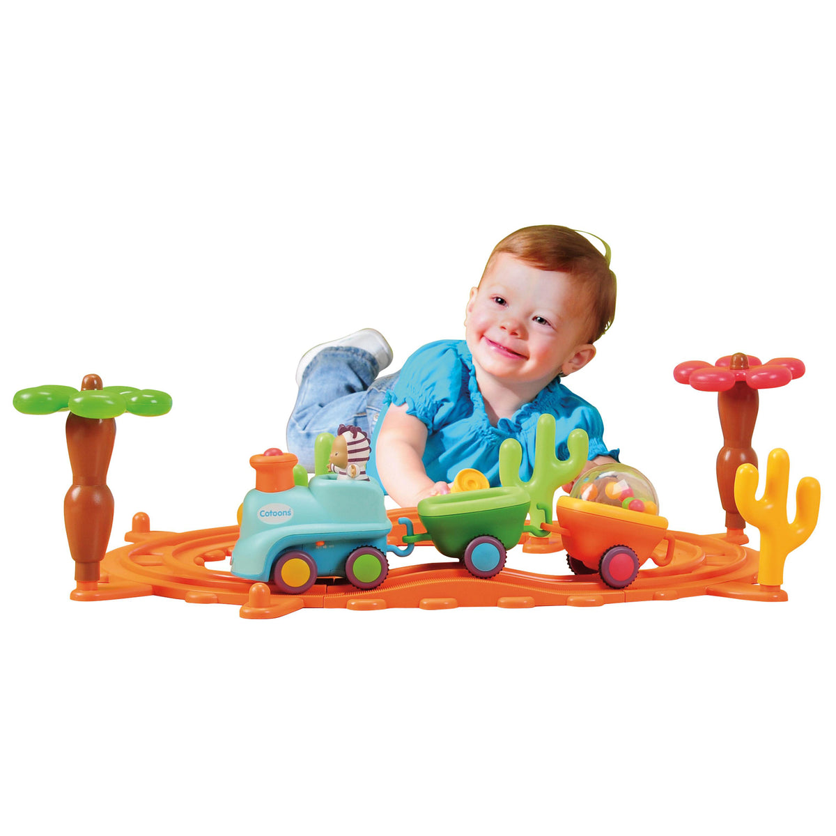 A smiling toddler lies on an orange toy train track with a small turquoise train, green cargo car, yellow cactus, and flower-shaped track pieces on a white background.