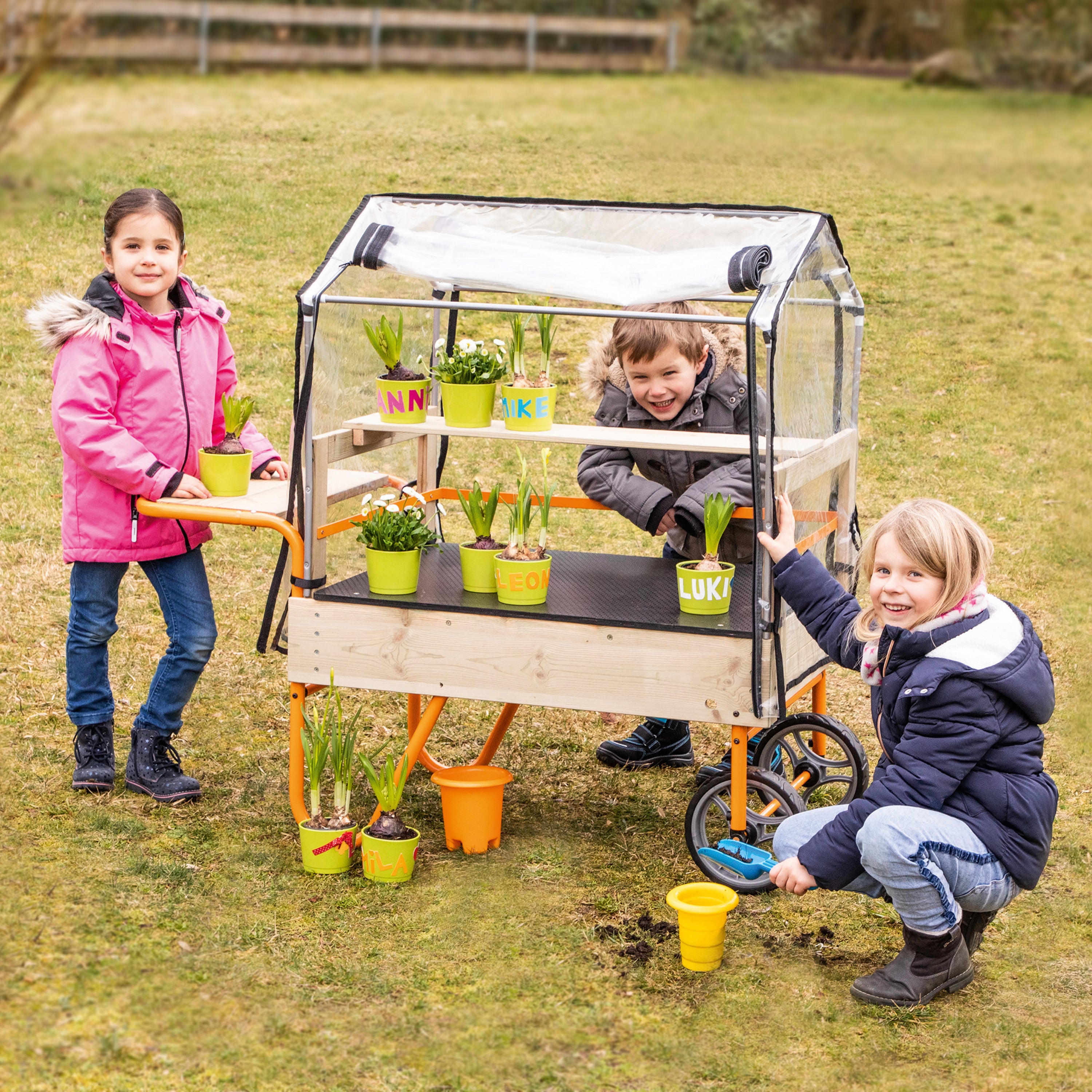Three children outdoors on a grassy lawn around a wooden garden cart with a clear plastic greenhouse cover, shelves holding small potted plants, and orange wheels.