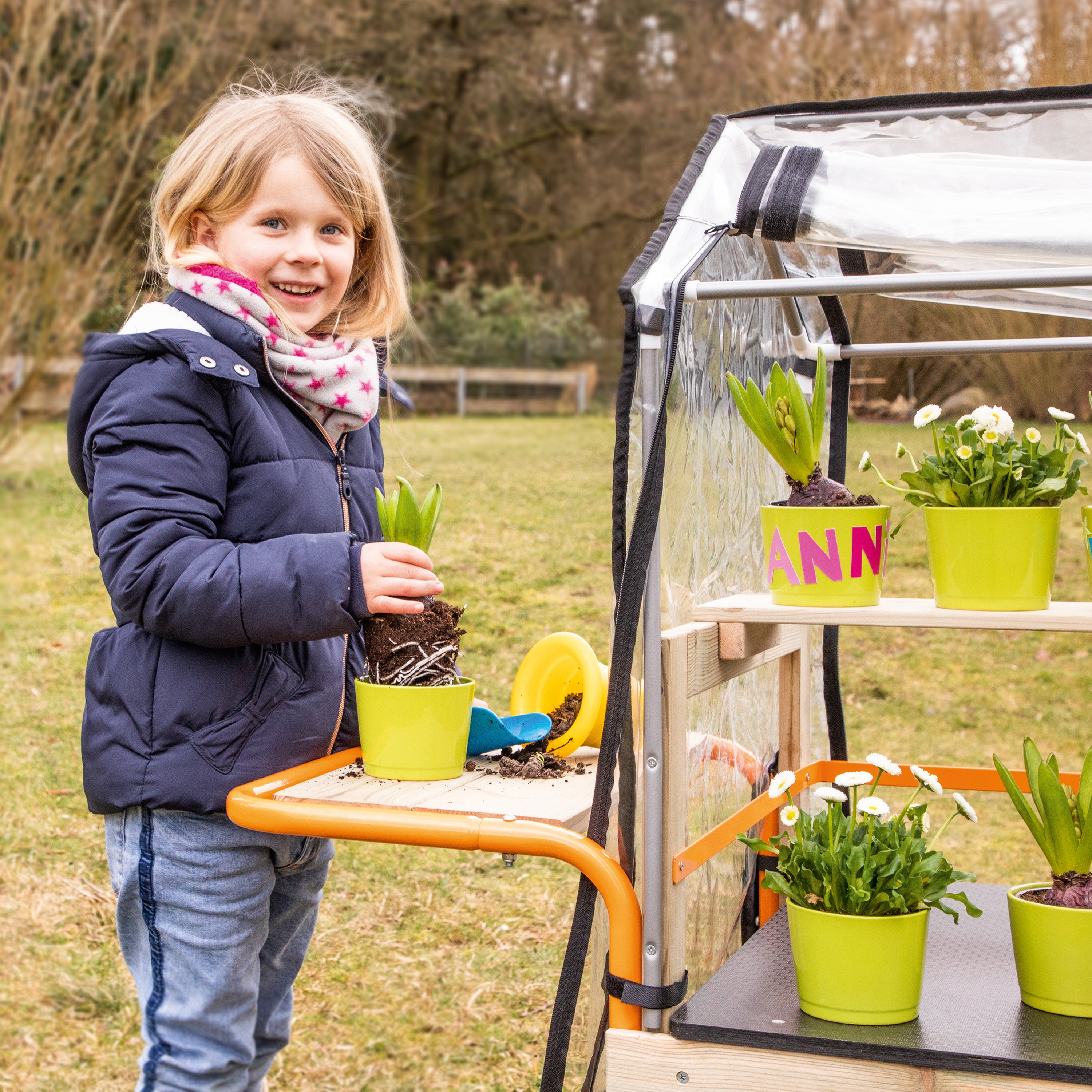 A young girl with blonde hair smiles while holding a small potted plant next to a wooden garden cart with a clear plastic cover. The cart has shelves holding several bright green pots with plants, set in an outdoor grassy yard.