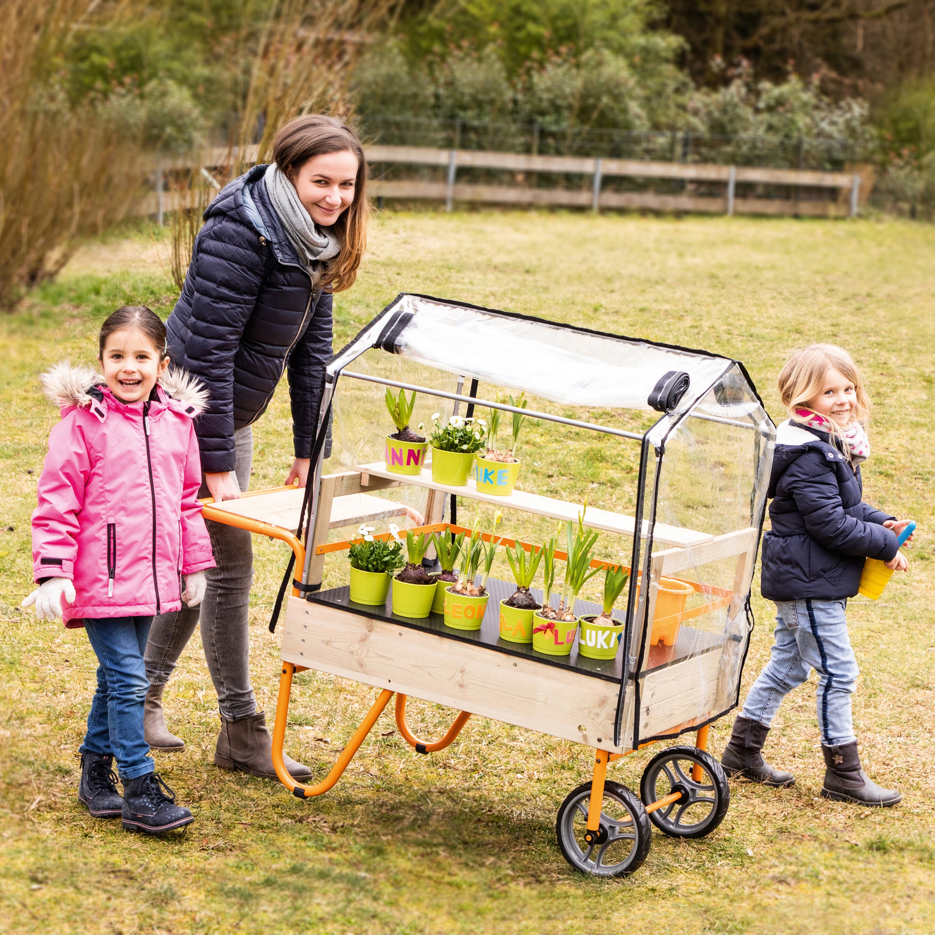 A woman and two children push a small wooden cart with a clear plastic greenhouse cover in an outdoor grassy park. The cart holds several small potted plants and flowers, and the children are dressed in winter jackets.