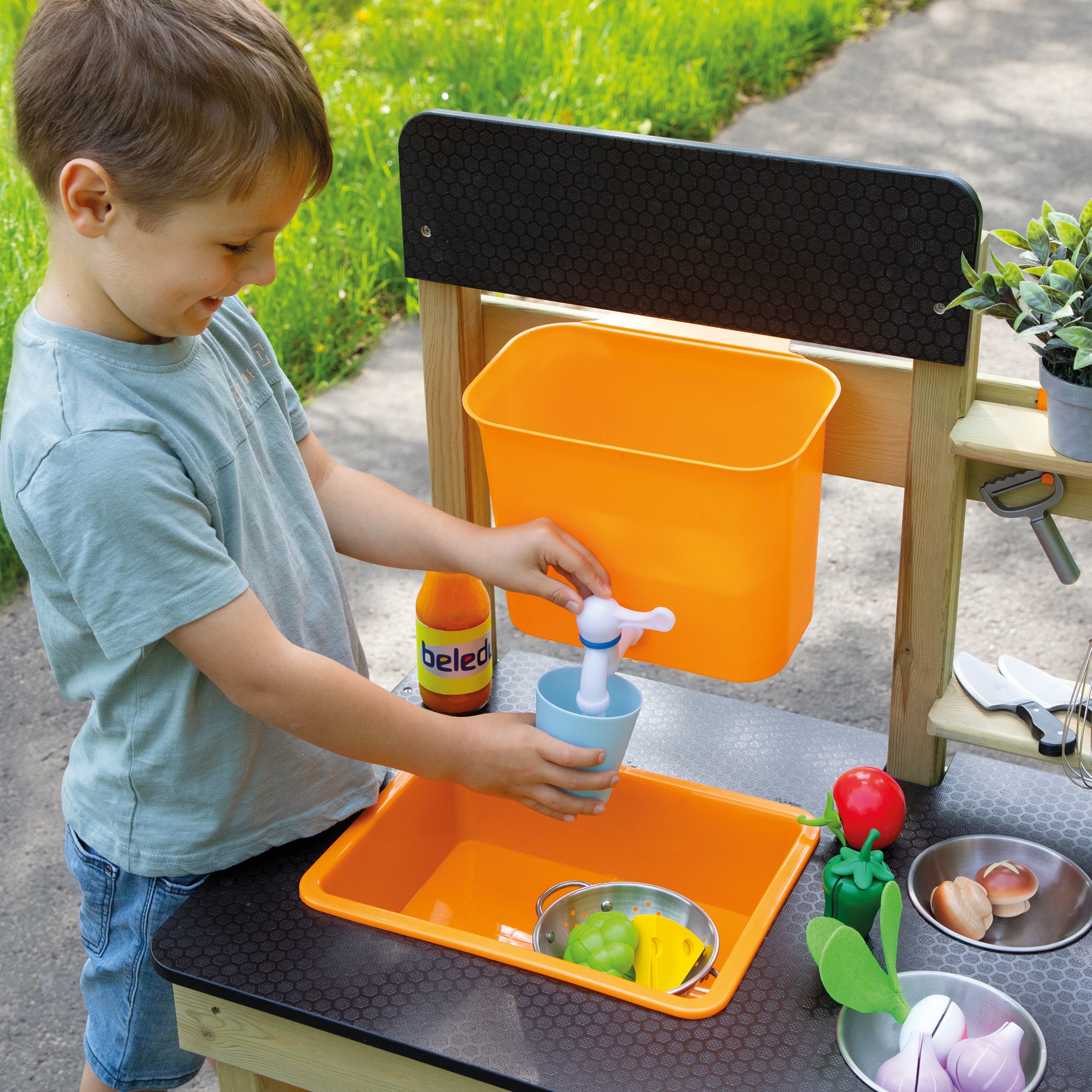A young boy playing with an outdoor wooden pretend play kitchen sink. He is washing a light blue cup under a black mesh backrest, using a toy faucet.