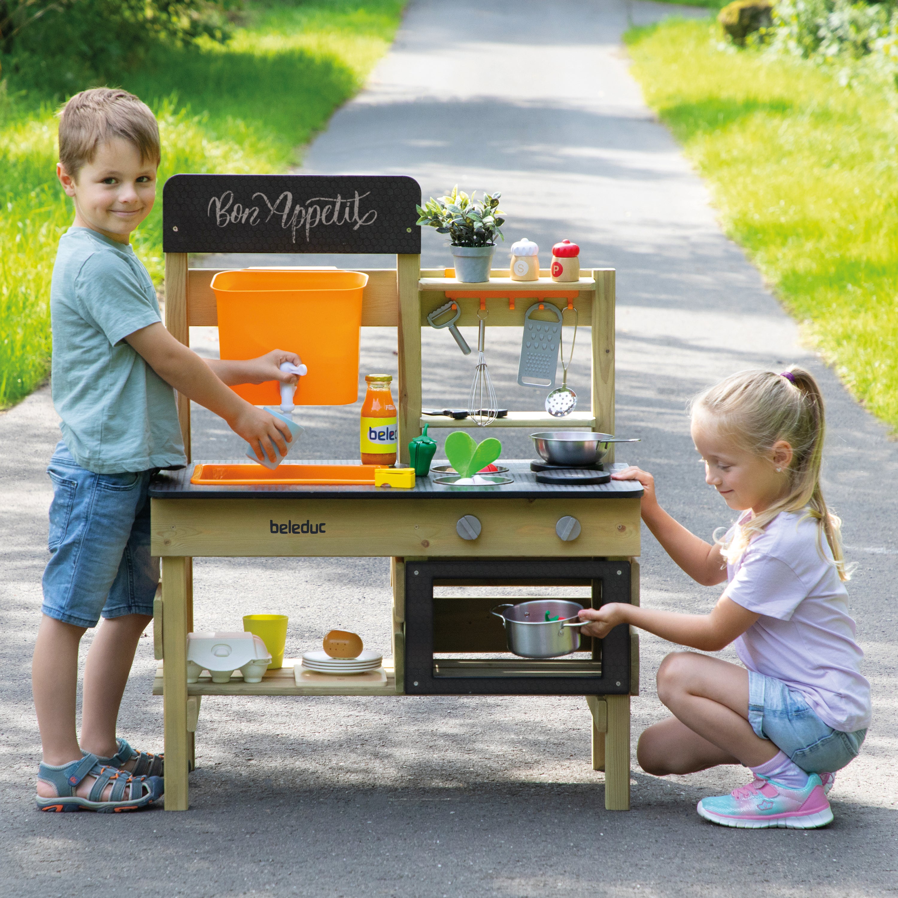 Two children playing with a wooden pretend kitchen playset outdoors on a paved path. The playset has a black front with orange, yellow, and green accessories, and the children are interacting with it in a sunny, grassy outdoor setting.