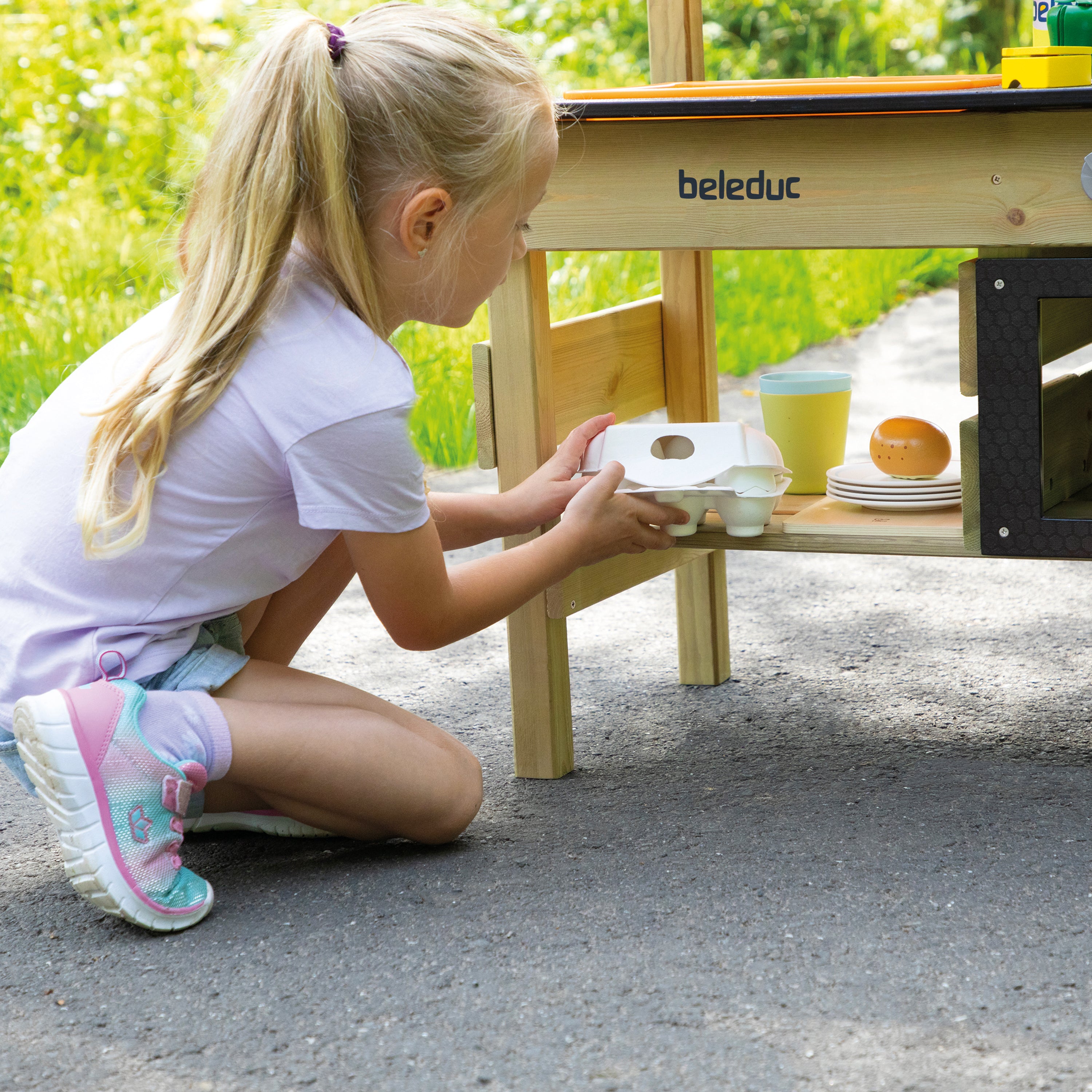 A young girl with blonde hair in a ponytail kneels on asphalt next to a wooden play kitchen table, placing a small white carton into the kitchen setting. The table has the brand name 'beleduc' printed on it.