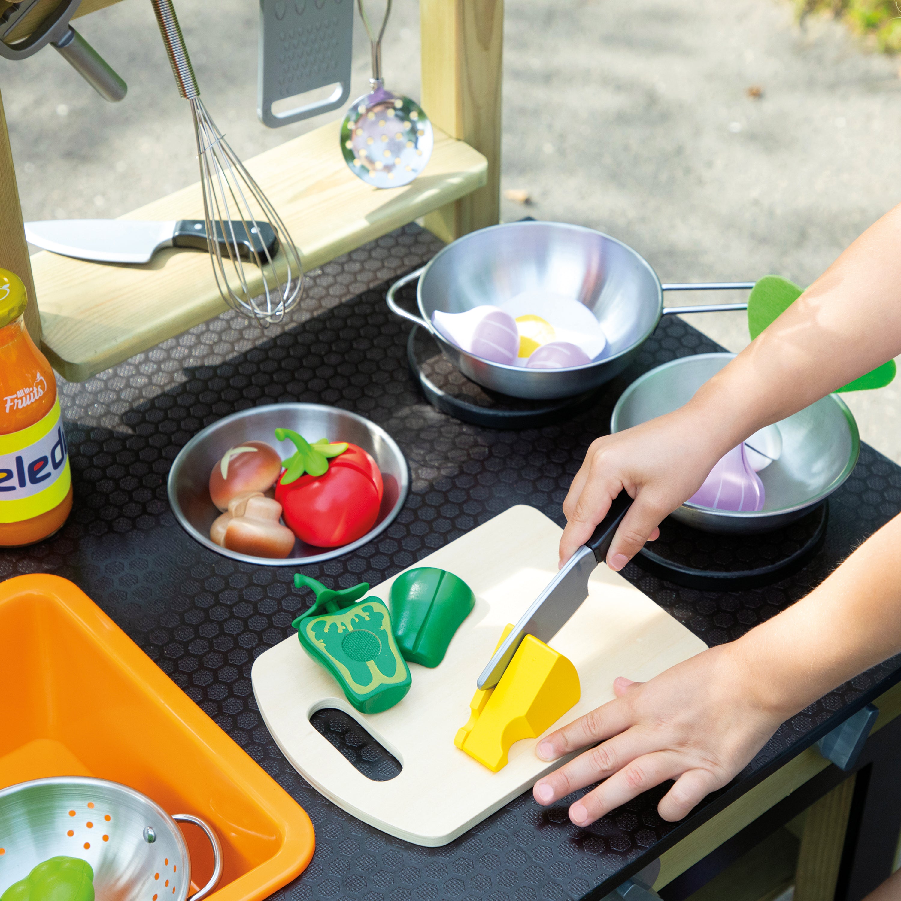 A child's play kitchen scene with a person's hands using a toy knife to cut a yellow toy vegetable on a cream cutting board. The play kitchen setup includes toy pots, a wooden shelf with hanging utensils, and colorful toy vegetables on a dark patterned countertop.