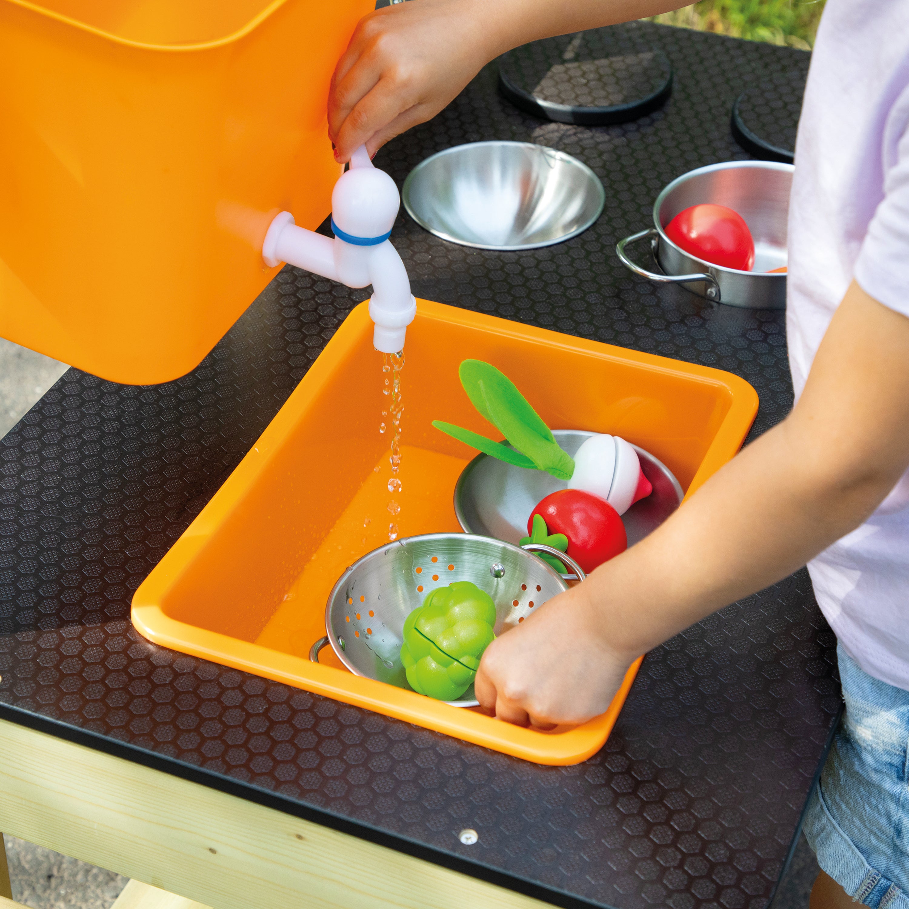 A child's hands wash colorful toy vegetables in an orange play sink with a faucet, with an orange bucket pouring water. The scene is set on a black picnic table outdoors.