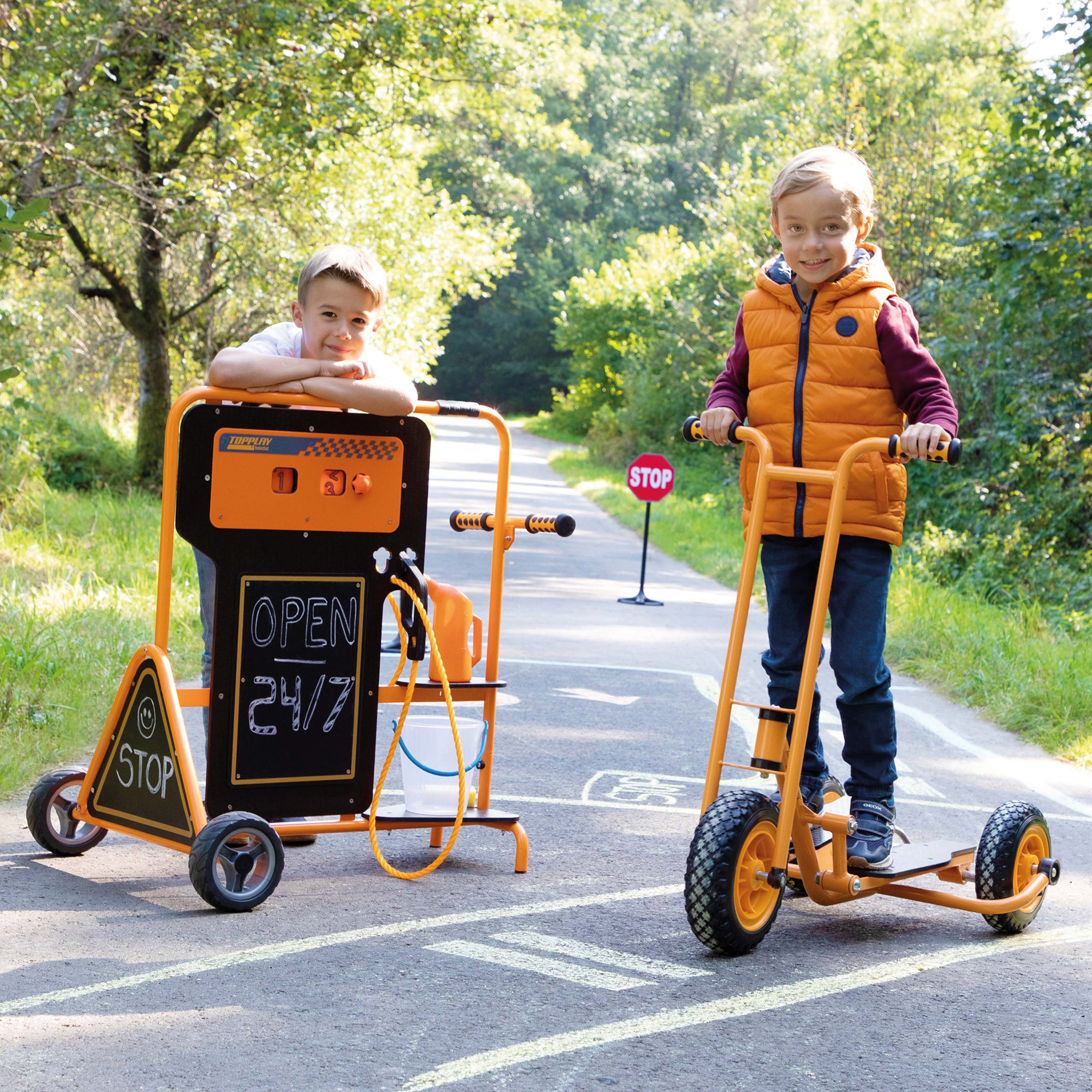 Two children outdoors on a paved path beside a grassy area. One child stands on an orange and black balance scooter, while the other leans on an orange and black utility cart with a chalkboard sign that reads 'OPEN 24/7'.