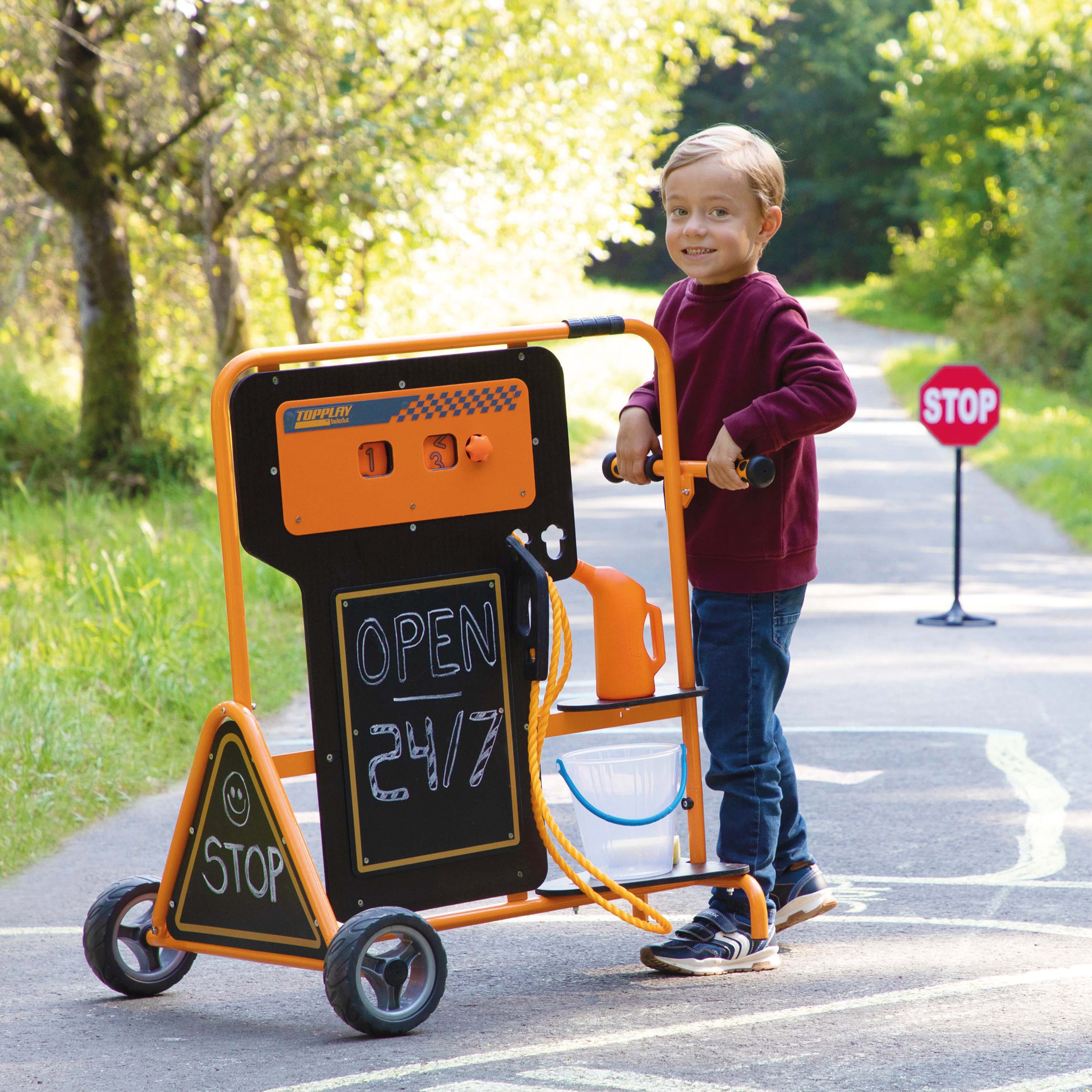 A young boy stands on an outdoor paved path behind a toy traffic light and stop sign cart. The toy cart has an orange frame, black sign panels, wheels, and small accessories, with the boy holding the handlebar.
