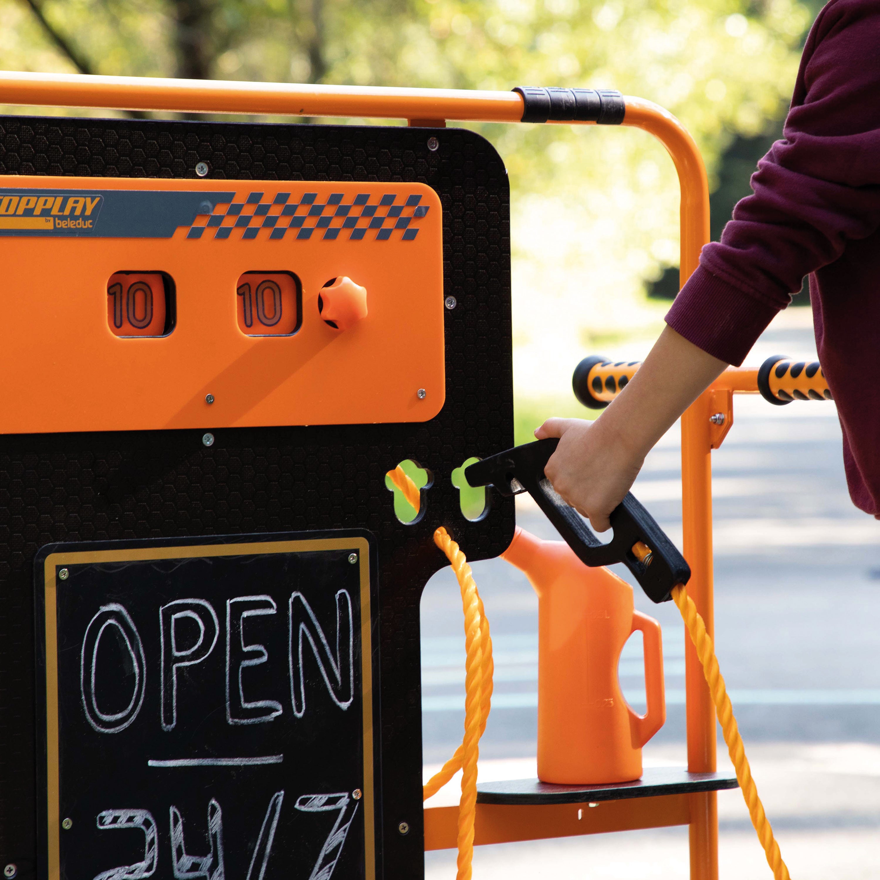 A person in a maroon sweatshirt uses a black spray nozzle on an outdoor playground water play structure. The structure is black and bright orange with a chalkboard sign reading 'OPEN 24/7' and a small orange pitcher on a shelf.