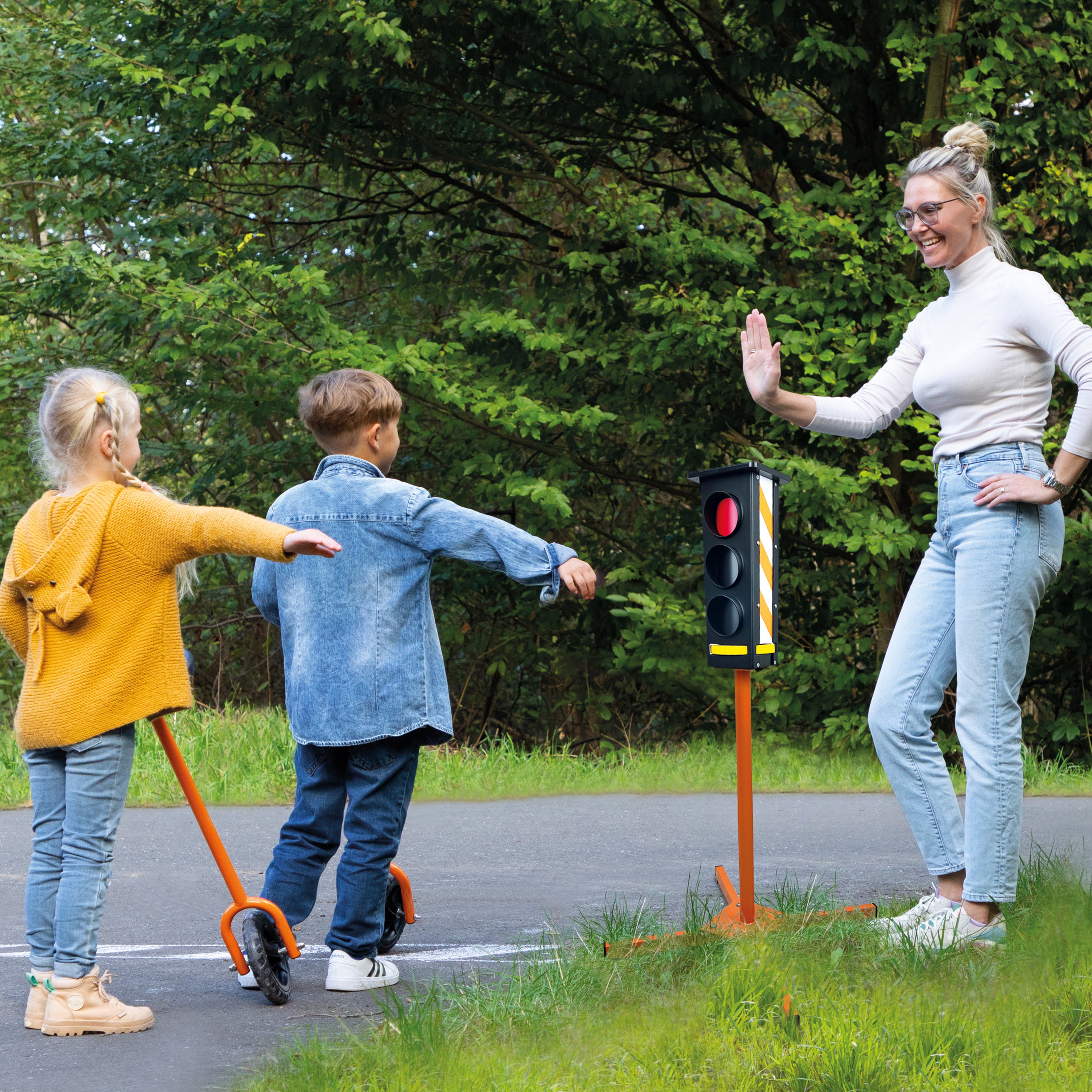A woman and two children outdoors near a small orange traffic light stand on a paved path. The woman is wearing a white long-sleeve top and light blue jeans, standing beside the stand and gesturing.