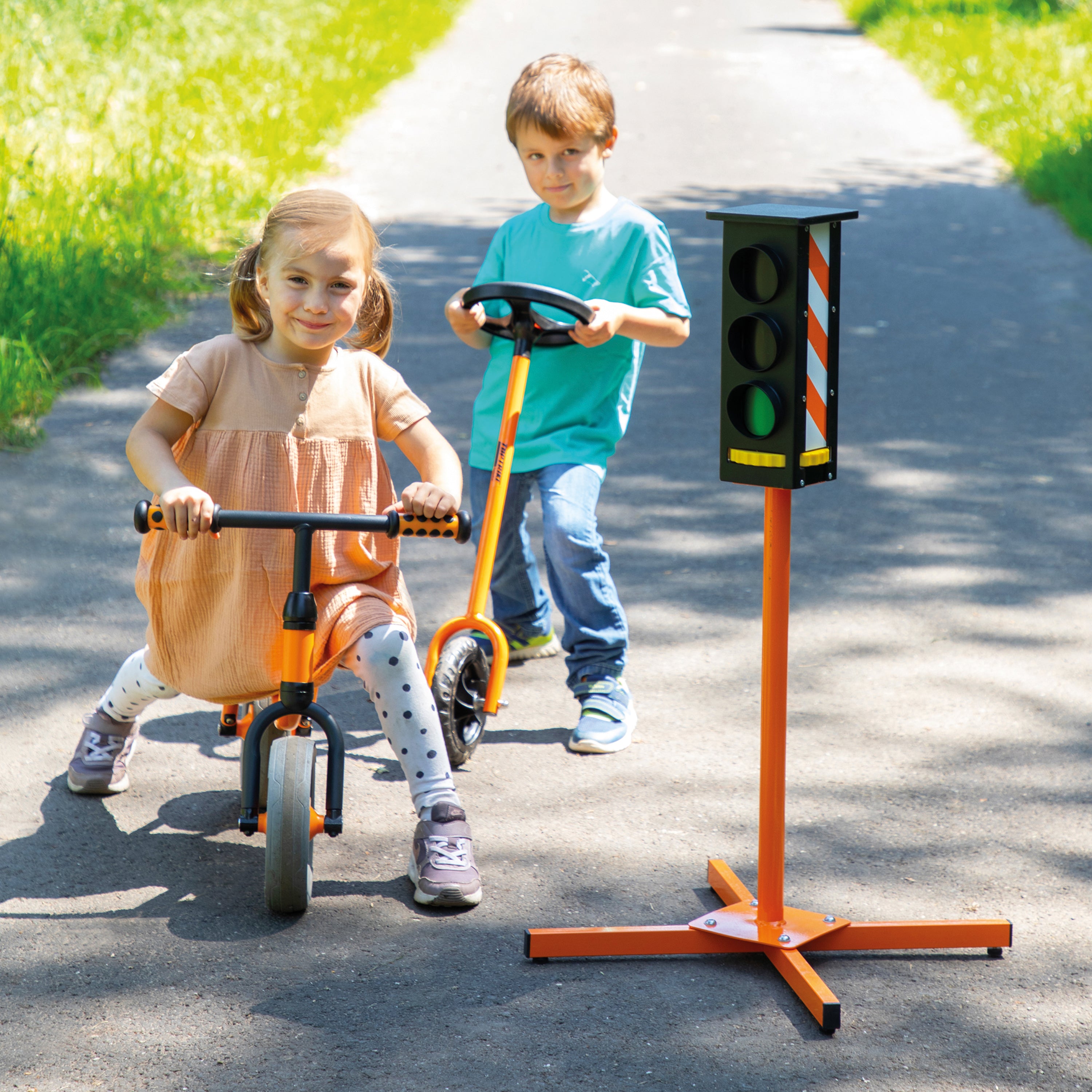Two children outdoors on a paved path with grass on both sides. One child rides a small orange balance bike, and the other pushes a matching orange toy scooter.