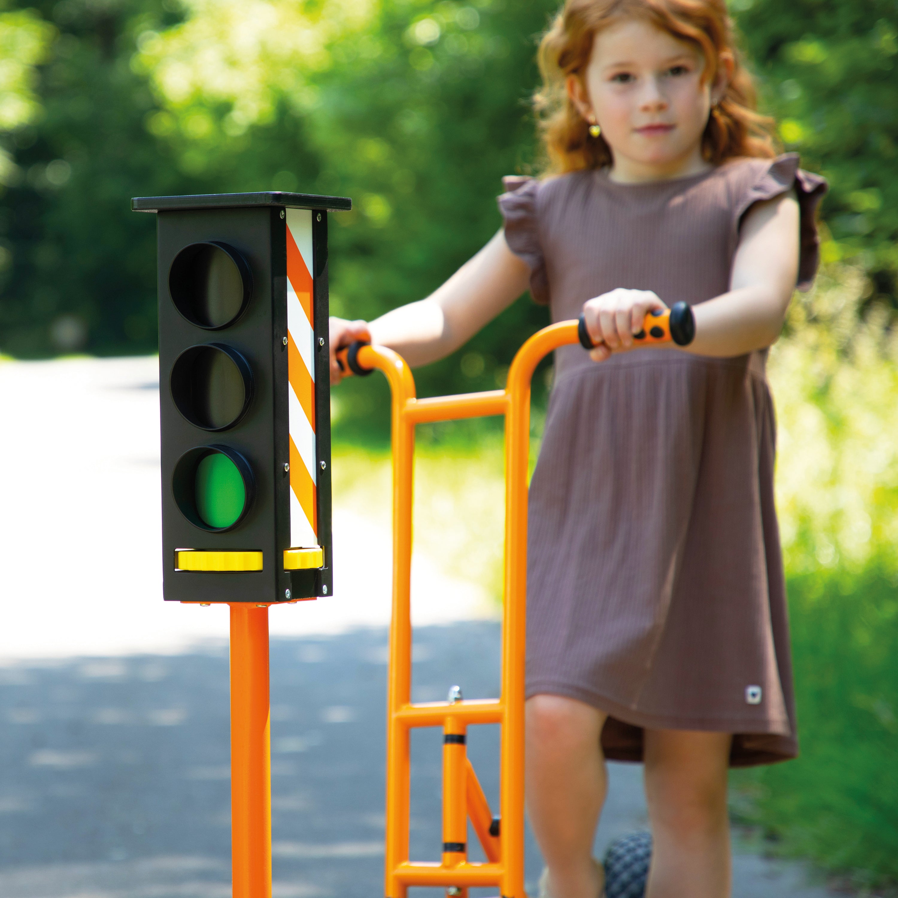 A young girl in a brown dress stands next to an orange playground traffic light toy with a ladder, holding the handlebar on an outdoor paved path with green trees in the background.