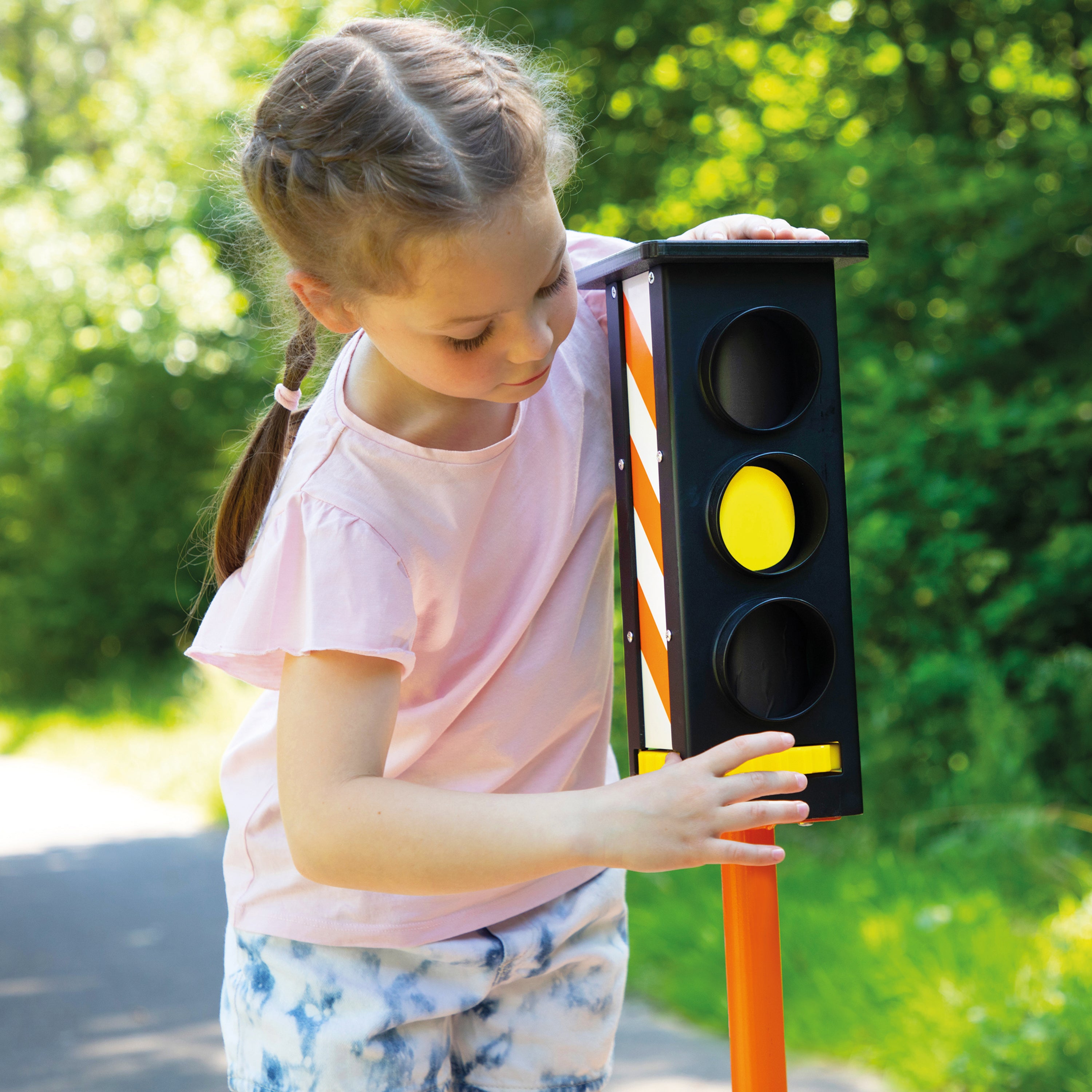 A young girl with braided hair stands outdoors on a path next to a toy traffic light with a black body, orange-and-white diagonal stripes, and a yellow center light. She is wearing a light pink t-shirt and blue-and-white patterned shorts.