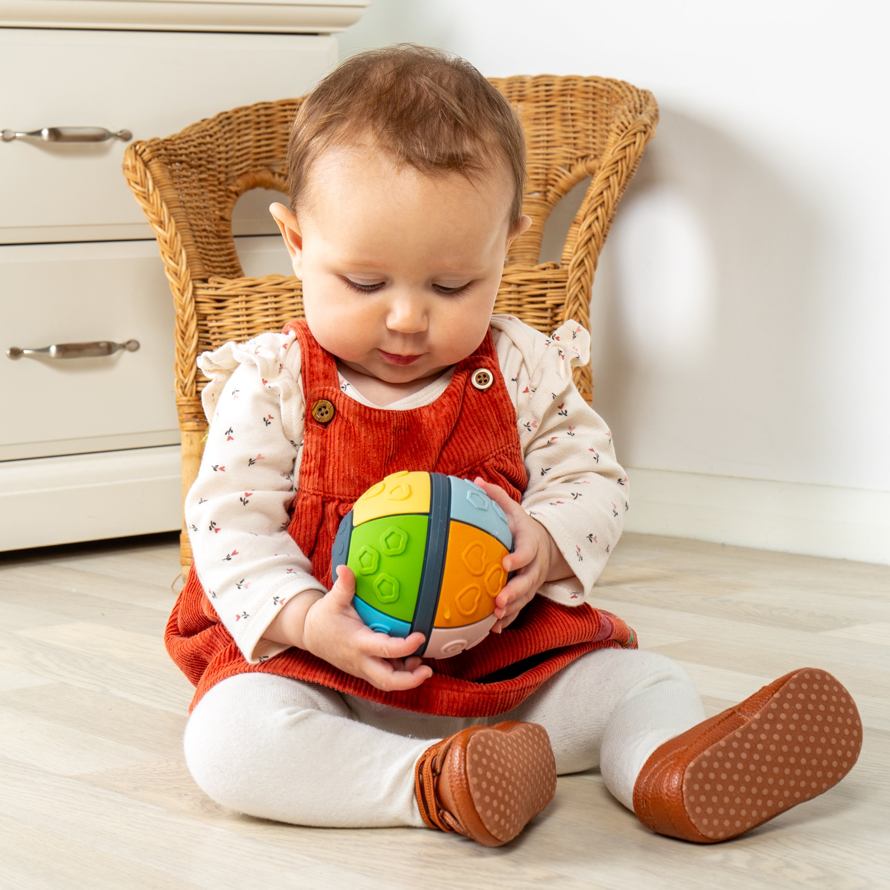 A baby sitting on a light wood floor in front of a wicker chair, holding a colorful, textured spherical toy. The baby is wearing a red corduroy pinafore dress over a white long-sleeve shirt with tiny red prints, white leggings, and brown moccasin-style shoes.