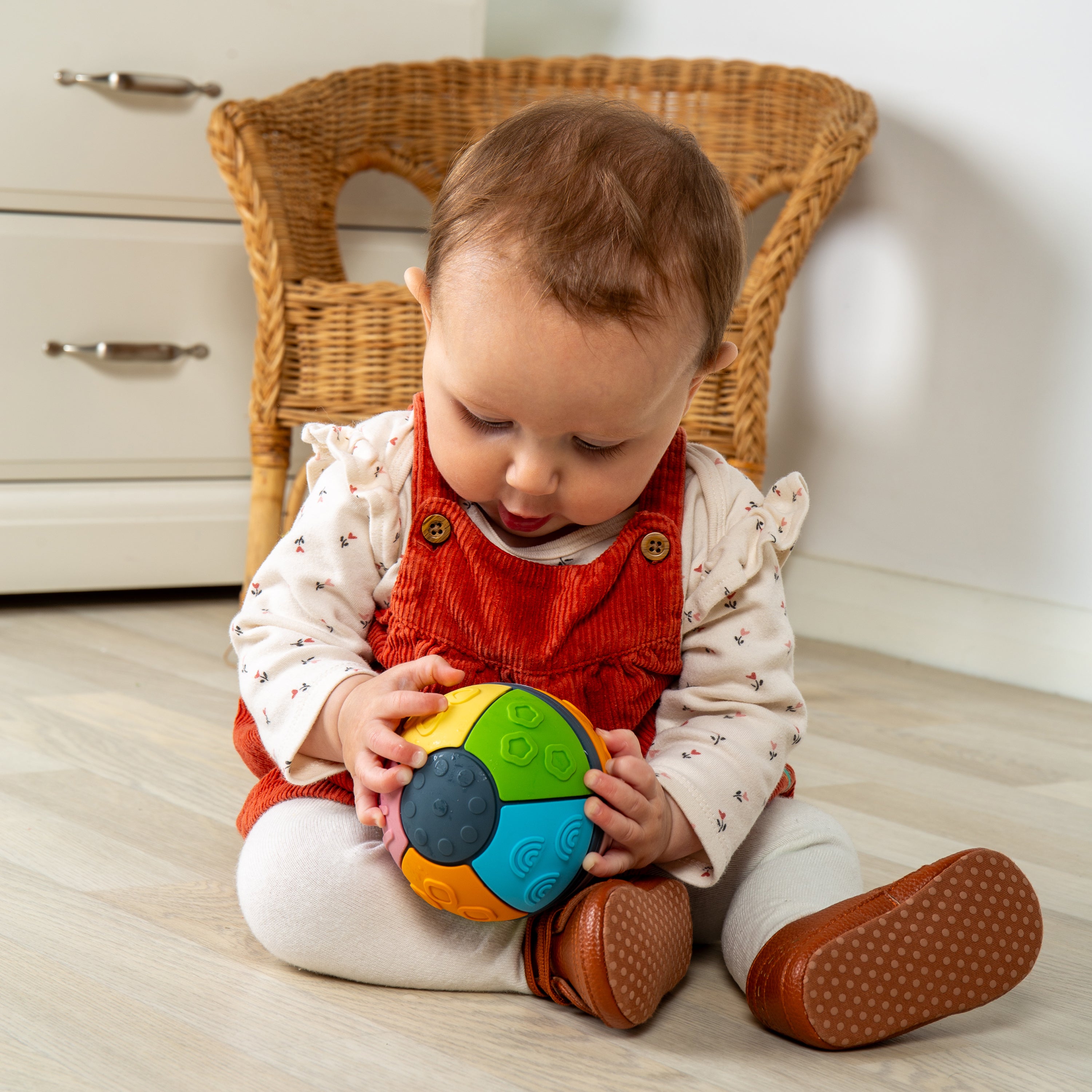 A baby sitting on a light wood floor in front of a wicker chair, holding a colorful segmented ball toy. The baby is wearing a red corduroy romper over a cream patterned long-sleeve shirt, white leggings, and brown shoes.