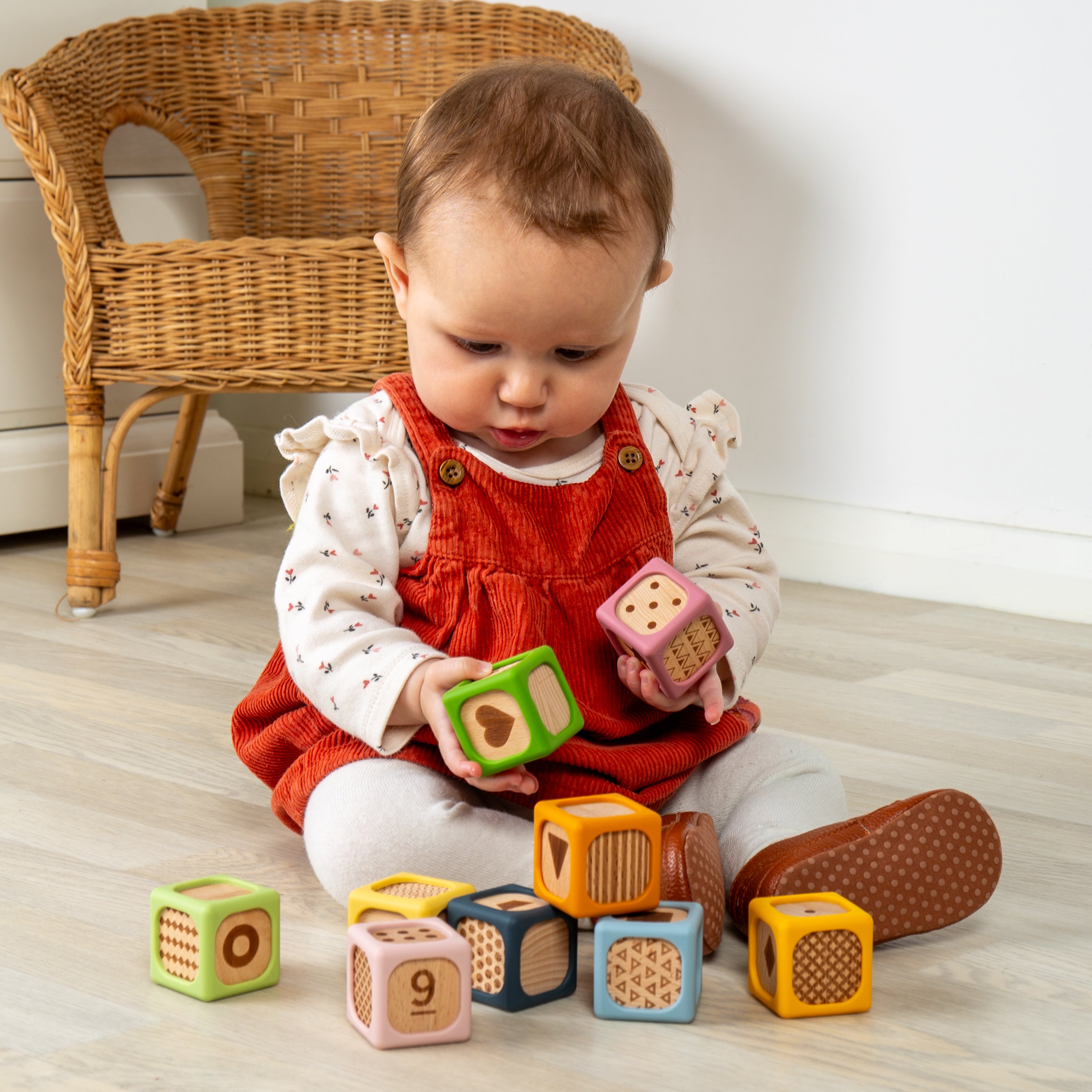 A baby sitting on a light wood floor, holding colorful wooden number and shape blocks. The baby is wearing a red corduroy pinafore dress over a white long-sleeve top with tiny printed dots, white leggings, and brown shoes.