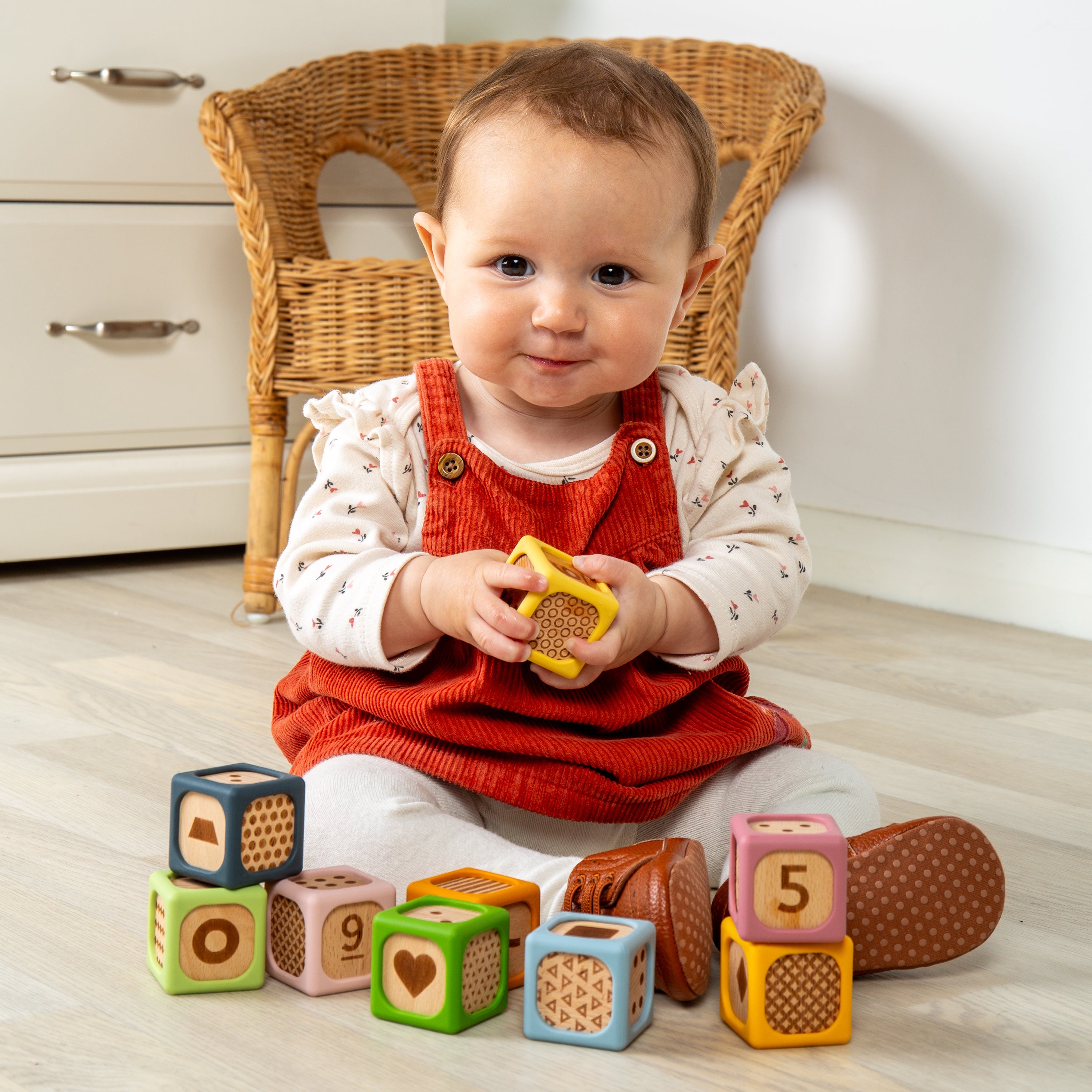 A baby sitting on a light wood floor in front of a wicker chair, holding a yellow toy while surrounded by colorful wooden number blocks.