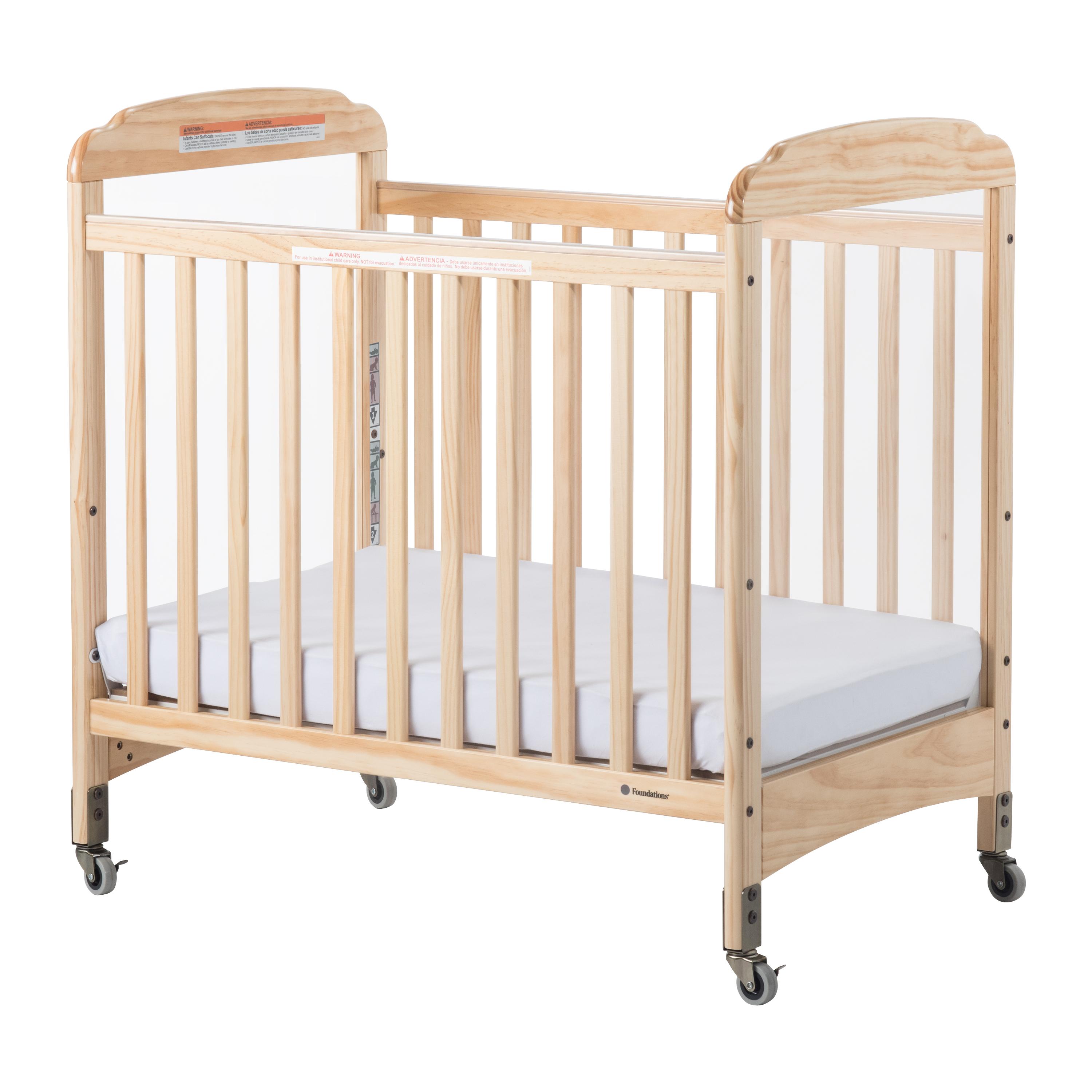 A light natural wood baby crib with a white mattress, shown on a clean white background. The crib features a guard rail, adjustable height rails, and casters at the bottom.