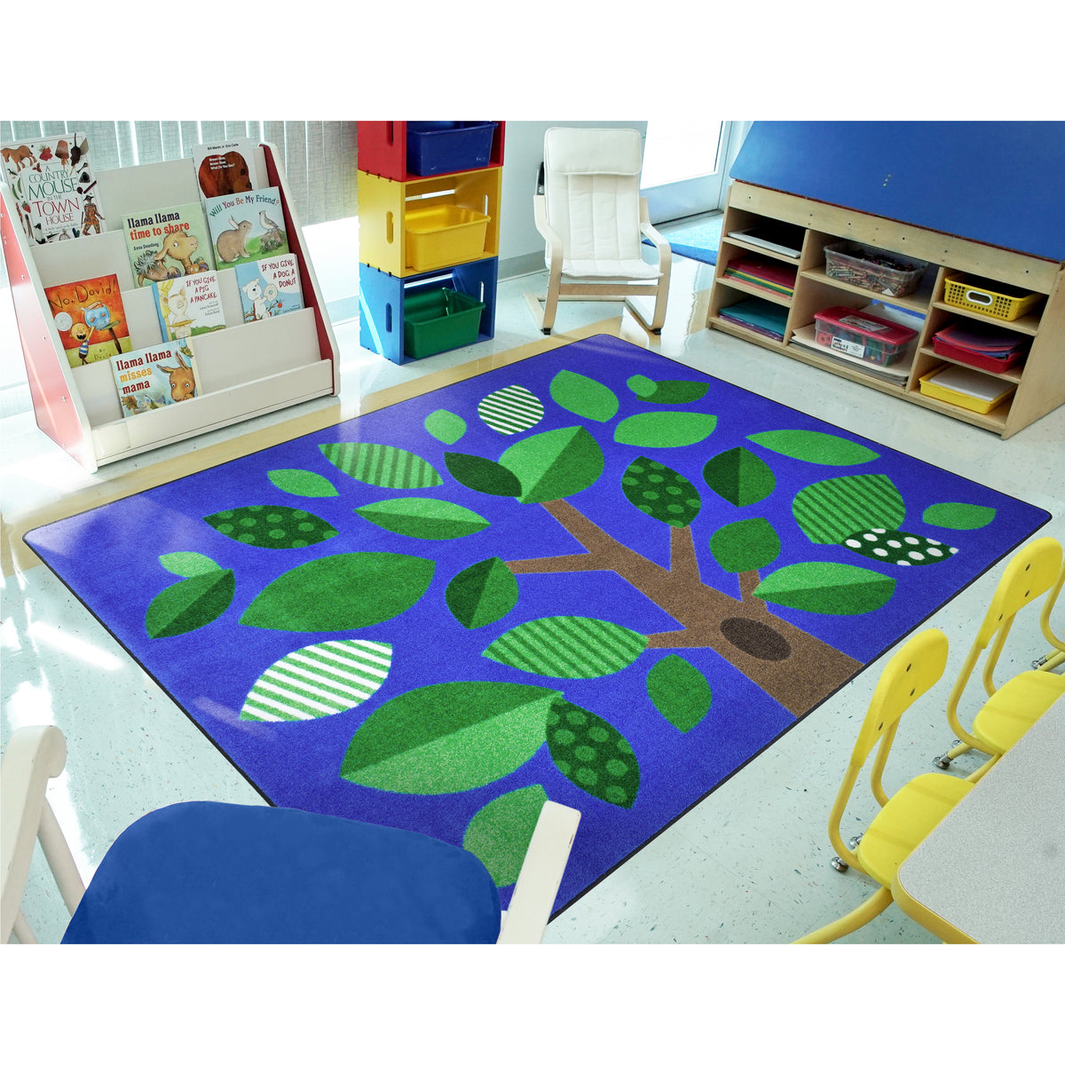 A colorful children's area rug with a stylized tree design in shades of green and brown on a bright blue-purple background, shown in a brightly lit preschool or daycare room with chairs, storage bins, and book displays.