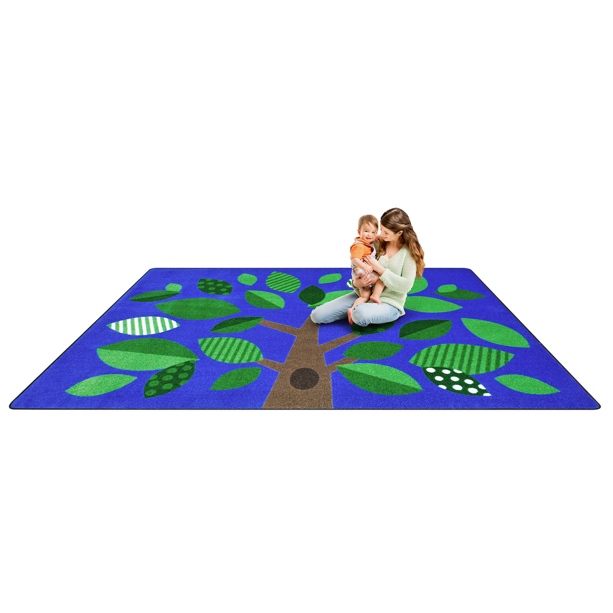 A large rectangular children's play rug featuring a stylized tree design with green leaves and a brown trunk on a blue background, shown with a woman and child sitting on it against a white studio background.