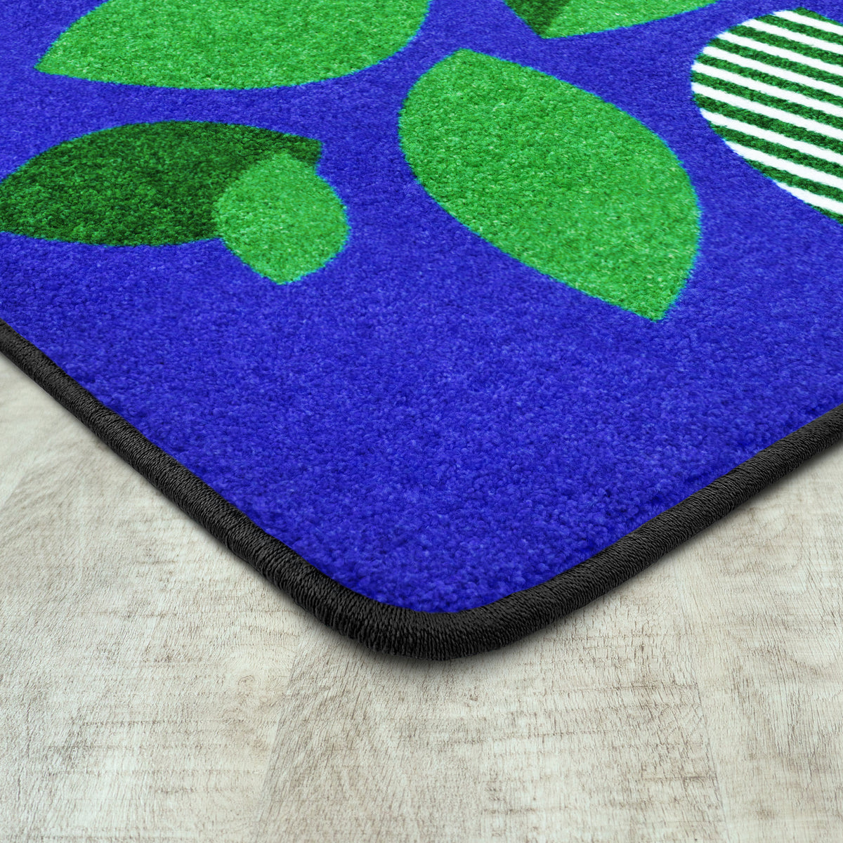 A close-up of a textured rug corner showing a bright blue base with large green leaf shapes, a green-and-white striped circular motif, and a black stitched border on a light wood floor.
