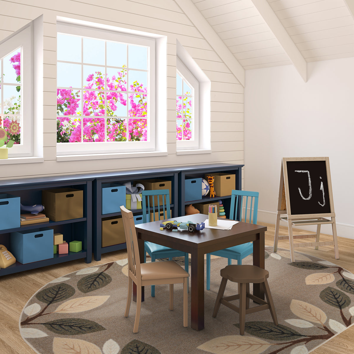 A styled children's playroom corner featuring a dark wood table, two teal chairs, a small stool, and a black chalkboard on an easel. The furniture is arranged on a leaf-patterned rug in front of built-in navy storage shelves with tan and blue bins, set against white shiplap walls and windows showing bright pink flowers outside.