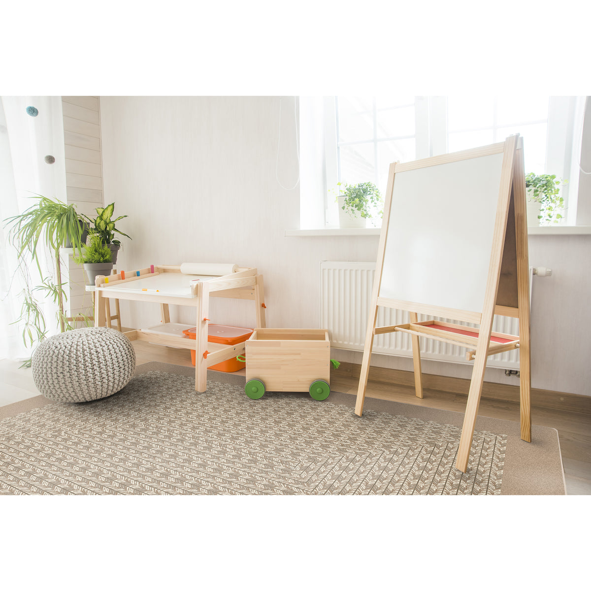 A children's wooden playroom set including a large whiteboard easel, a small wooden table, a wooden toy box with green wheels, and a gray knit pouf on a patterned rug in a bright room.