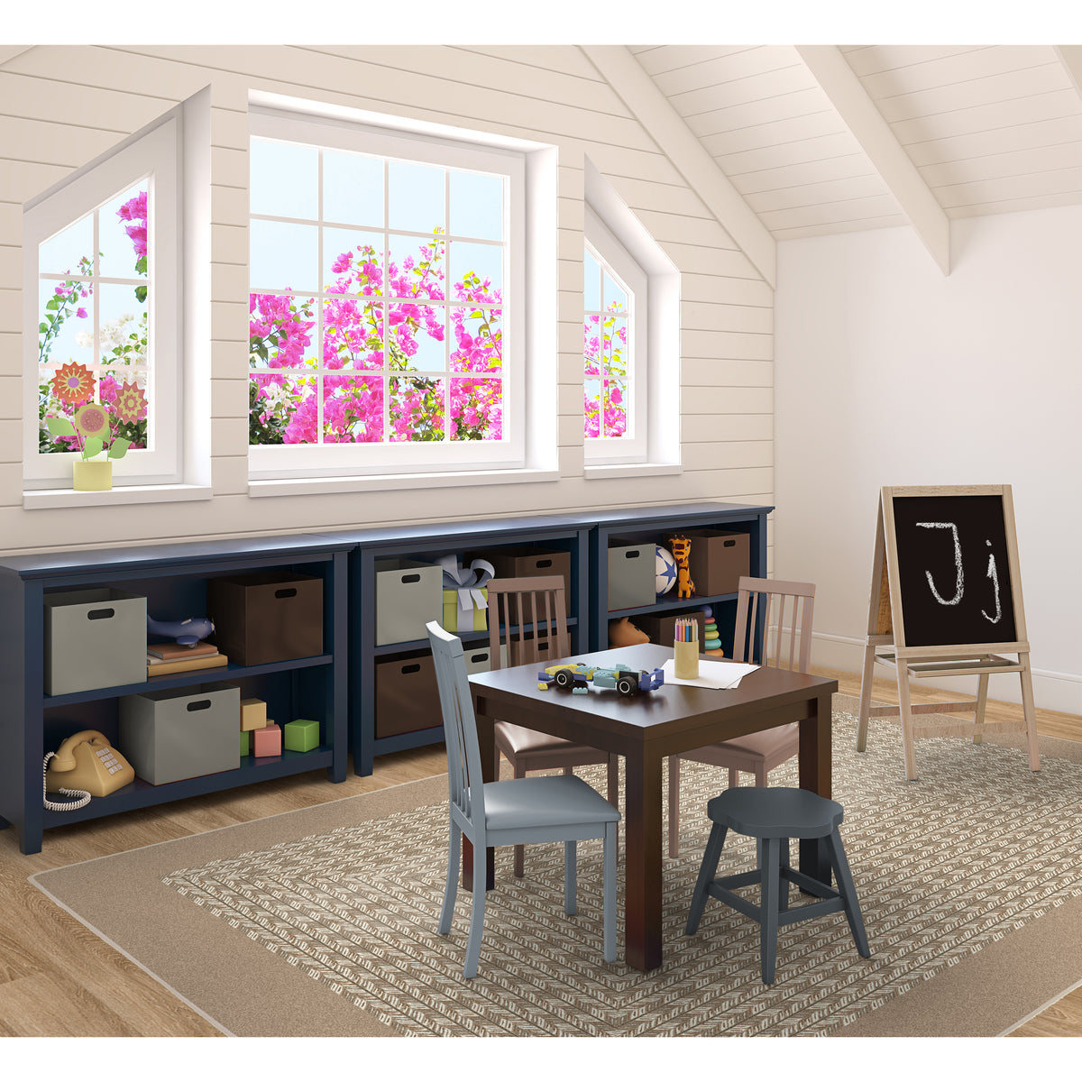 A styled children's playroom corner featuring a dark navy storage bench with bins, a wooden activity table with chairs and a small stool, a black chalkboard on a wooden easel, and a patterned area rug. Large windows with pink flowers are visible in the background.