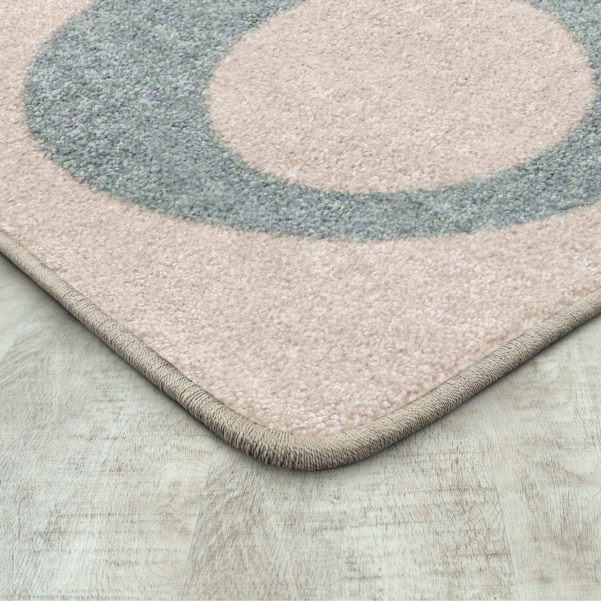 Close-up of a textured area rug corner showing a dusty pink field with a large muted blue-gray circular motif and a beige fringed edge on a light wood floor.