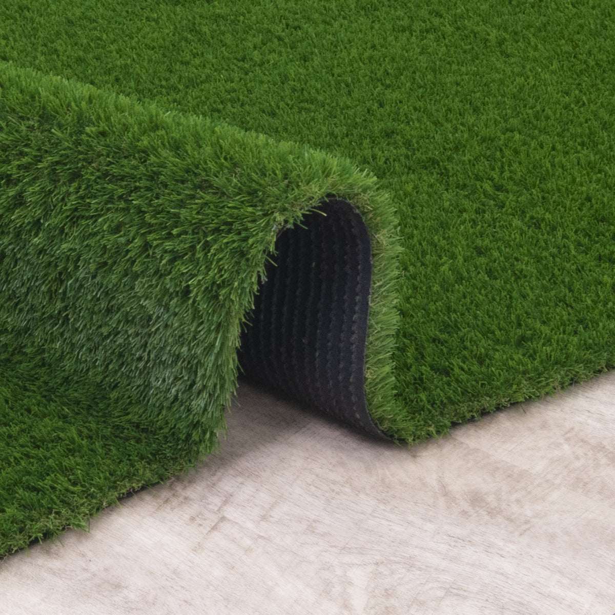 Green artificial turf or grass carpet shown rolled over to reveal a black backing, placed on a light beige stone or concrete surface.