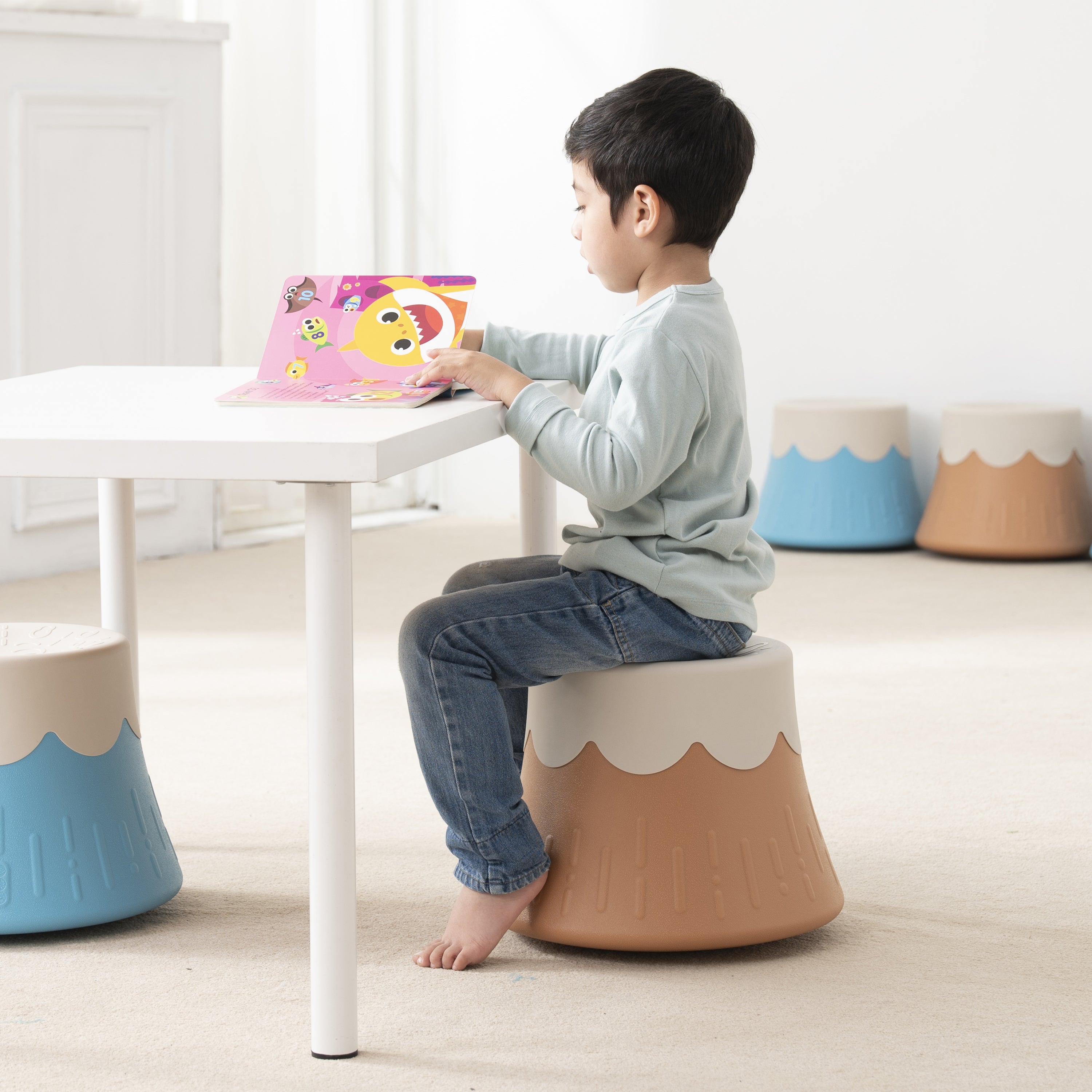 Child sitting on a small stool at a table with a colorful book.