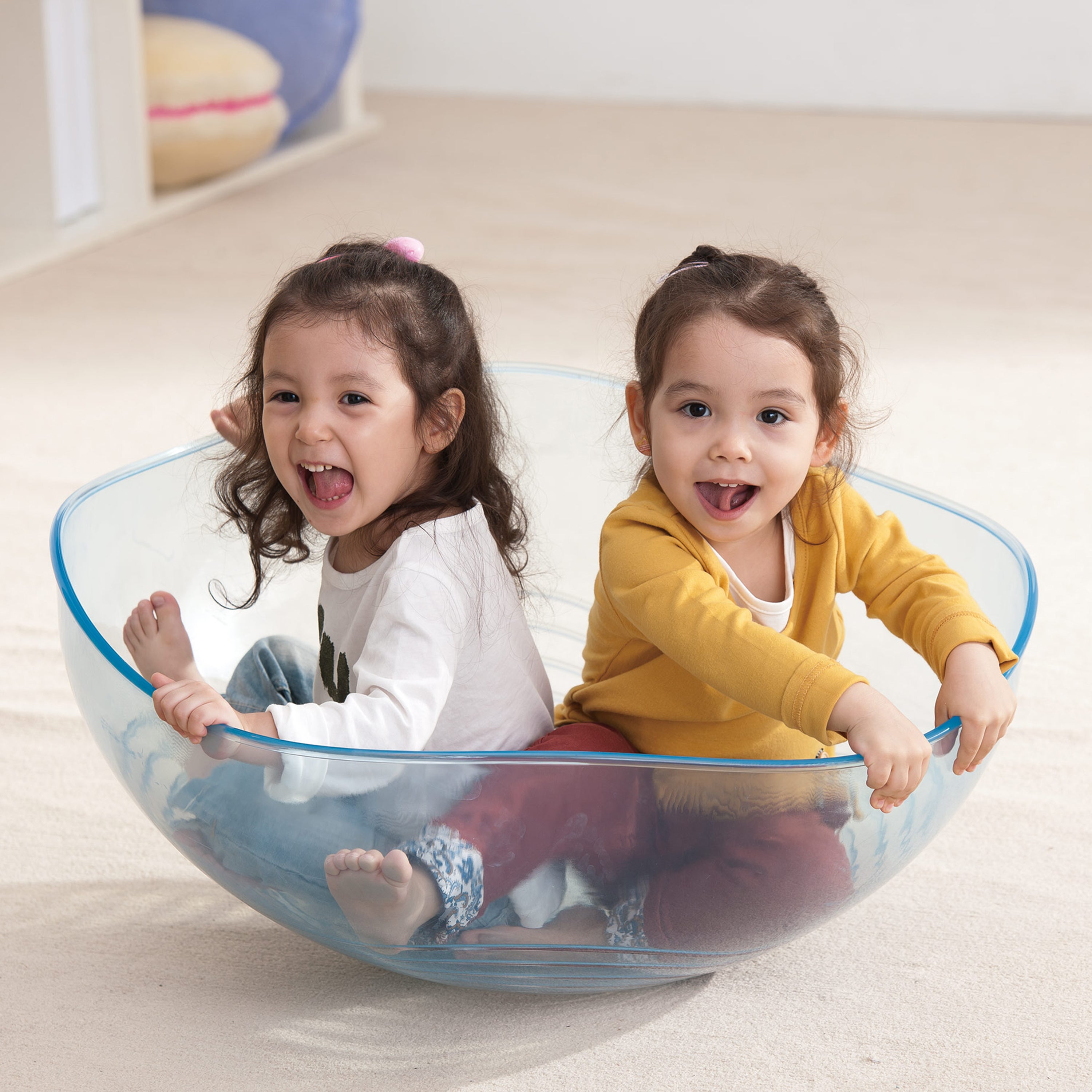 Two children playing inside a transparent plastic bowl on a light-colored floor.