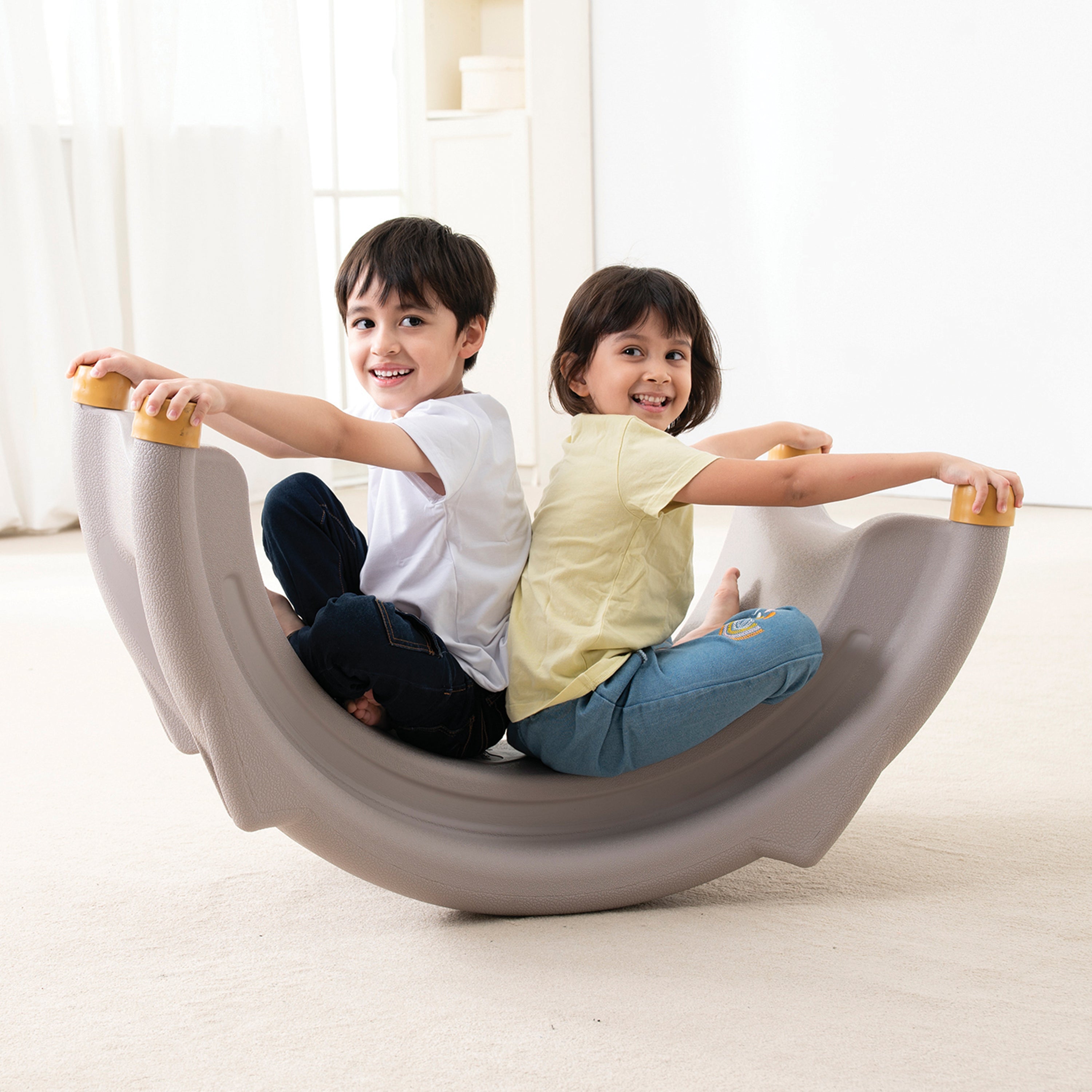 Two children sitting on a gray indoor seesaw in a bright room.