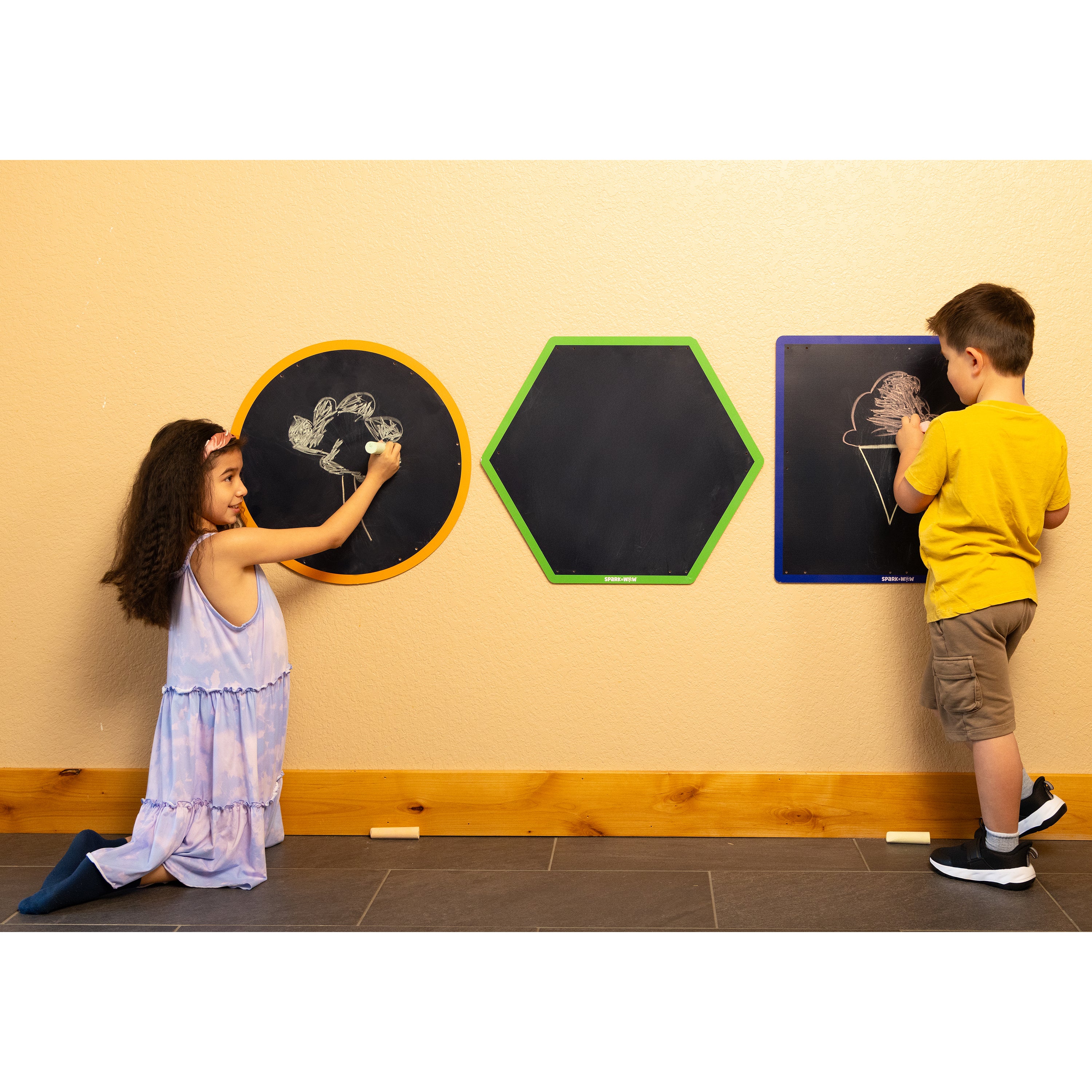 Two children drawing on hexagonal blackboards against a beige wall.