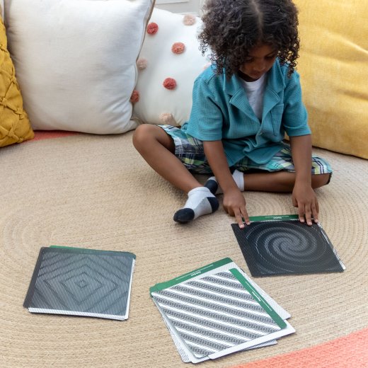 Child playing with geometric patterned cards on a carpeted floor.