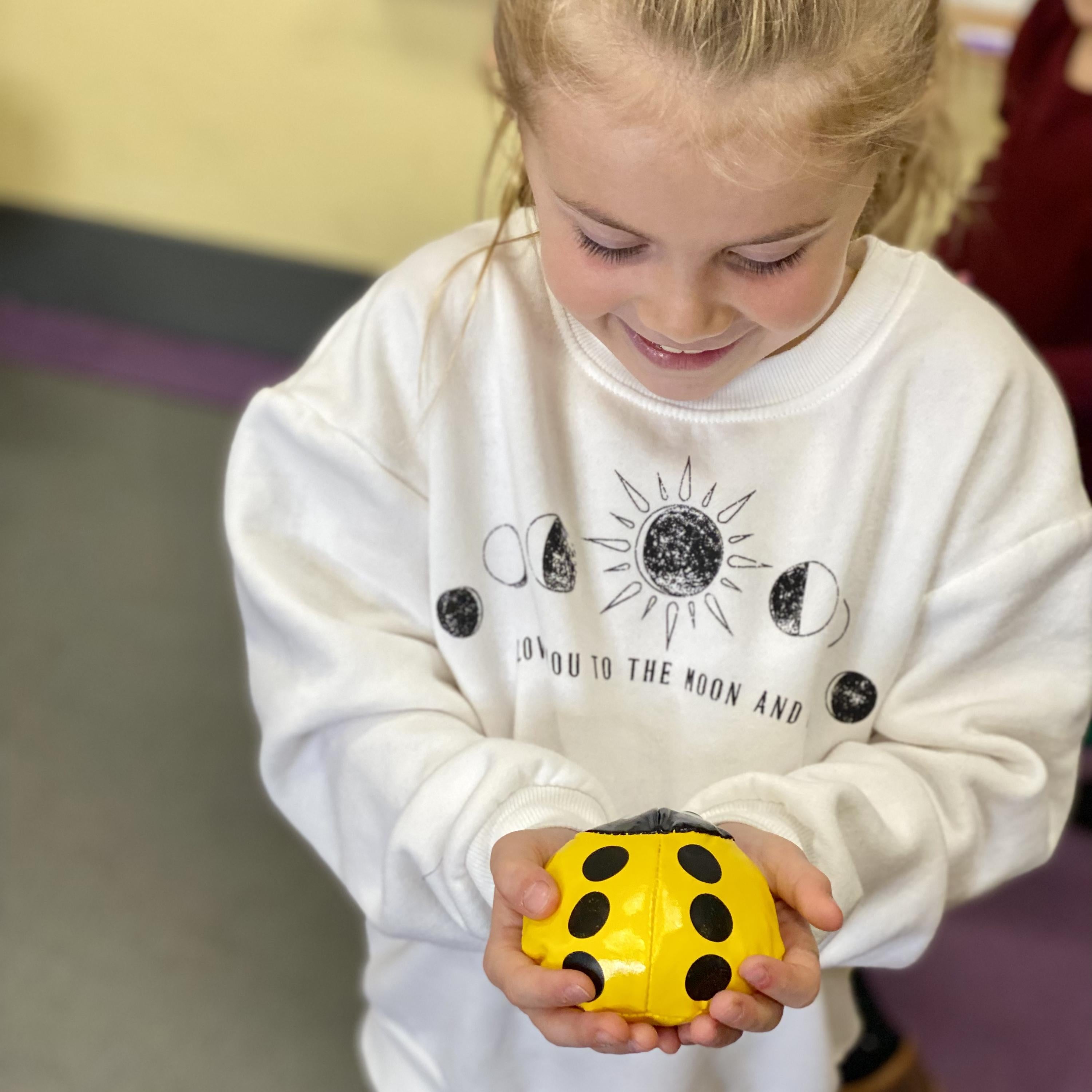 Child holding a yellow beanbag with black spots, wearing a white sweatshirt with graphics.