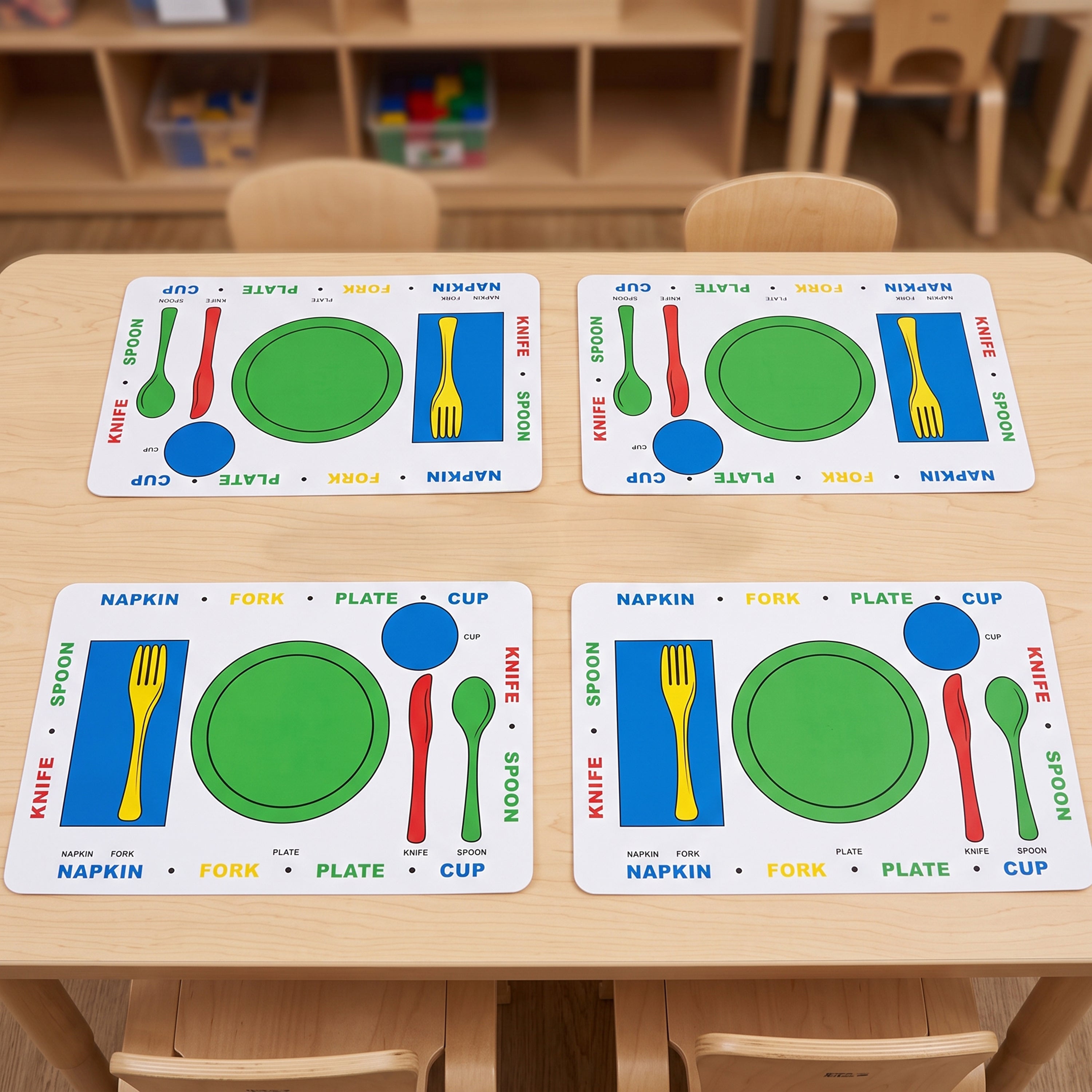 Four colorful children's table placemats arranged on a light wood table in a classroom setting. Each placemat shows labeled sections for knife, spoon, fork, plate, cup, and plate/cup with bright green, blue, red, yellow, and white colors.