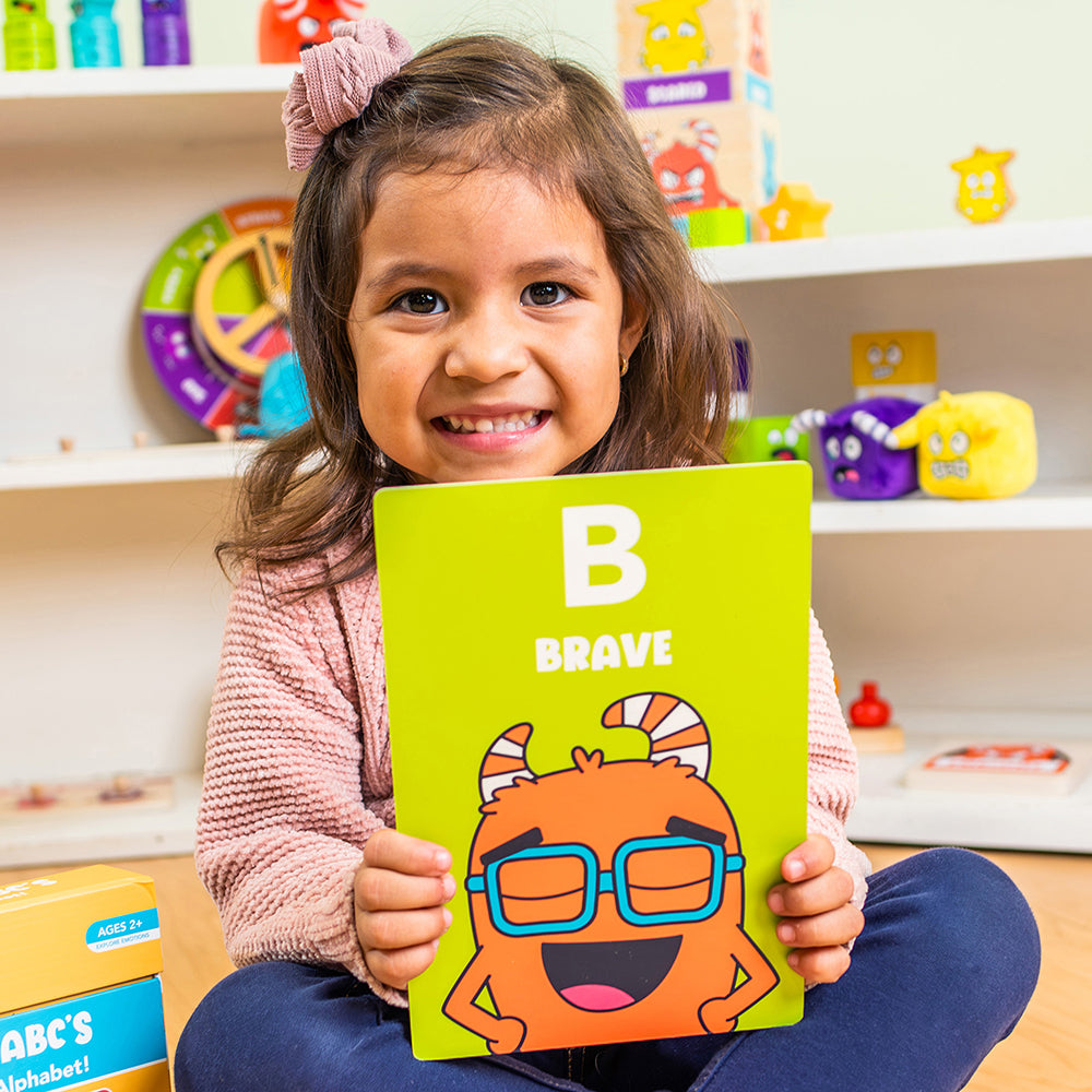 Child holding a jumbo card with the letter 'B' and the word 'Brave' in a colorful room.