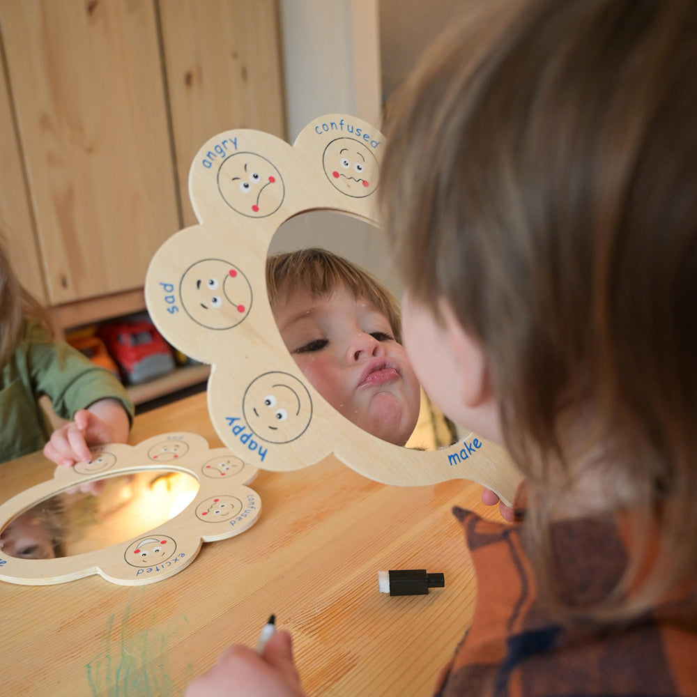 Child looking at a wooden mirror with emoticon faces and coloring it.