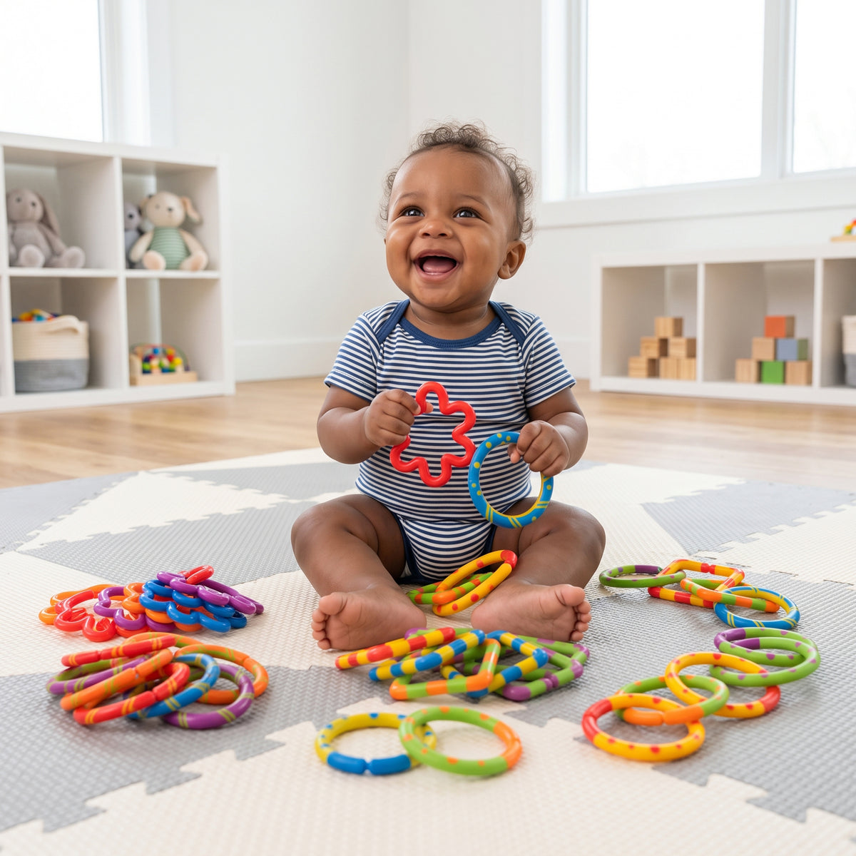A smiling baby sitting on a gray and white play mat in a bright nursery, holding colorful plastic ring toys while more of the same are scattered around them.