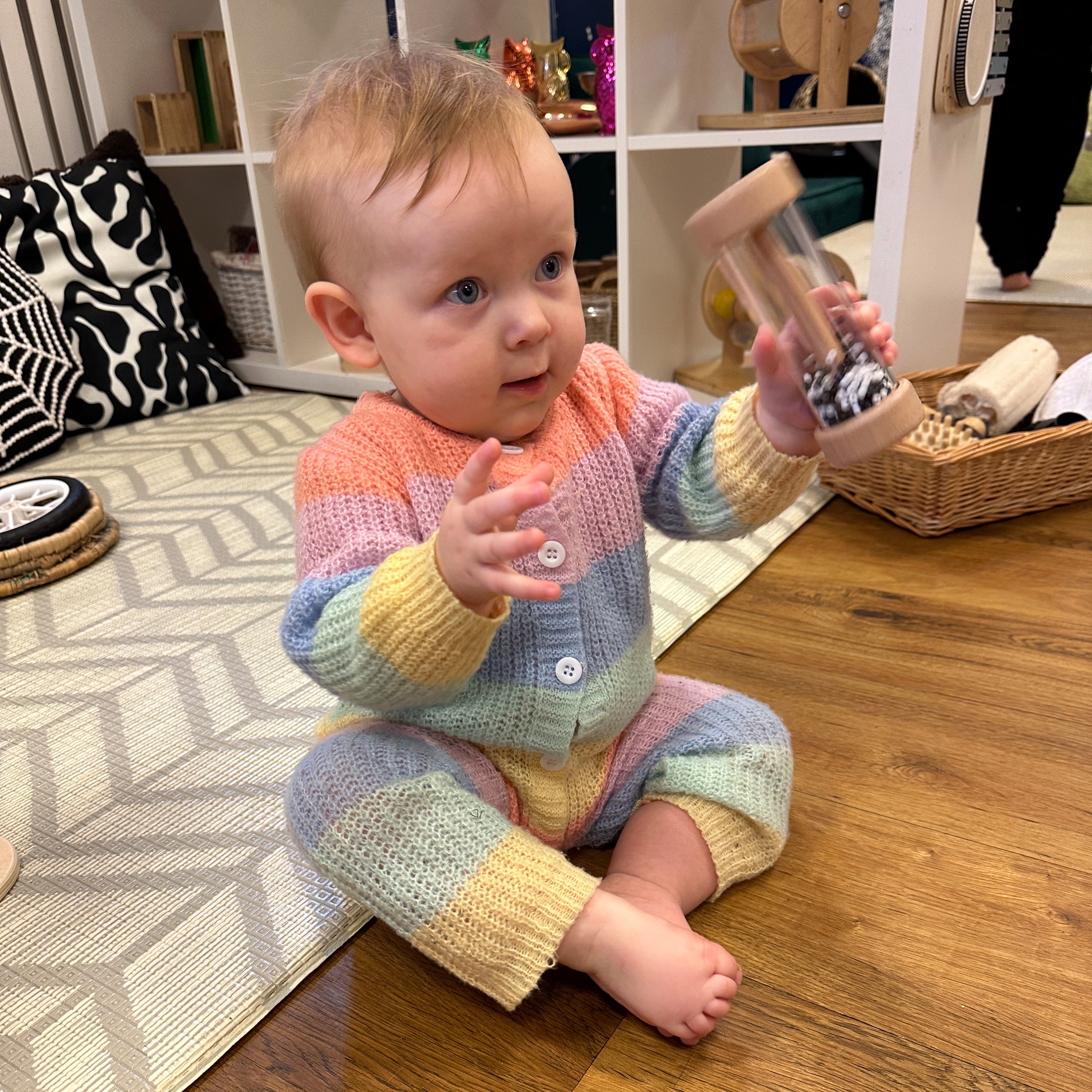 Baby in a colorful outfit sitting on a wooden floor holding a toy.