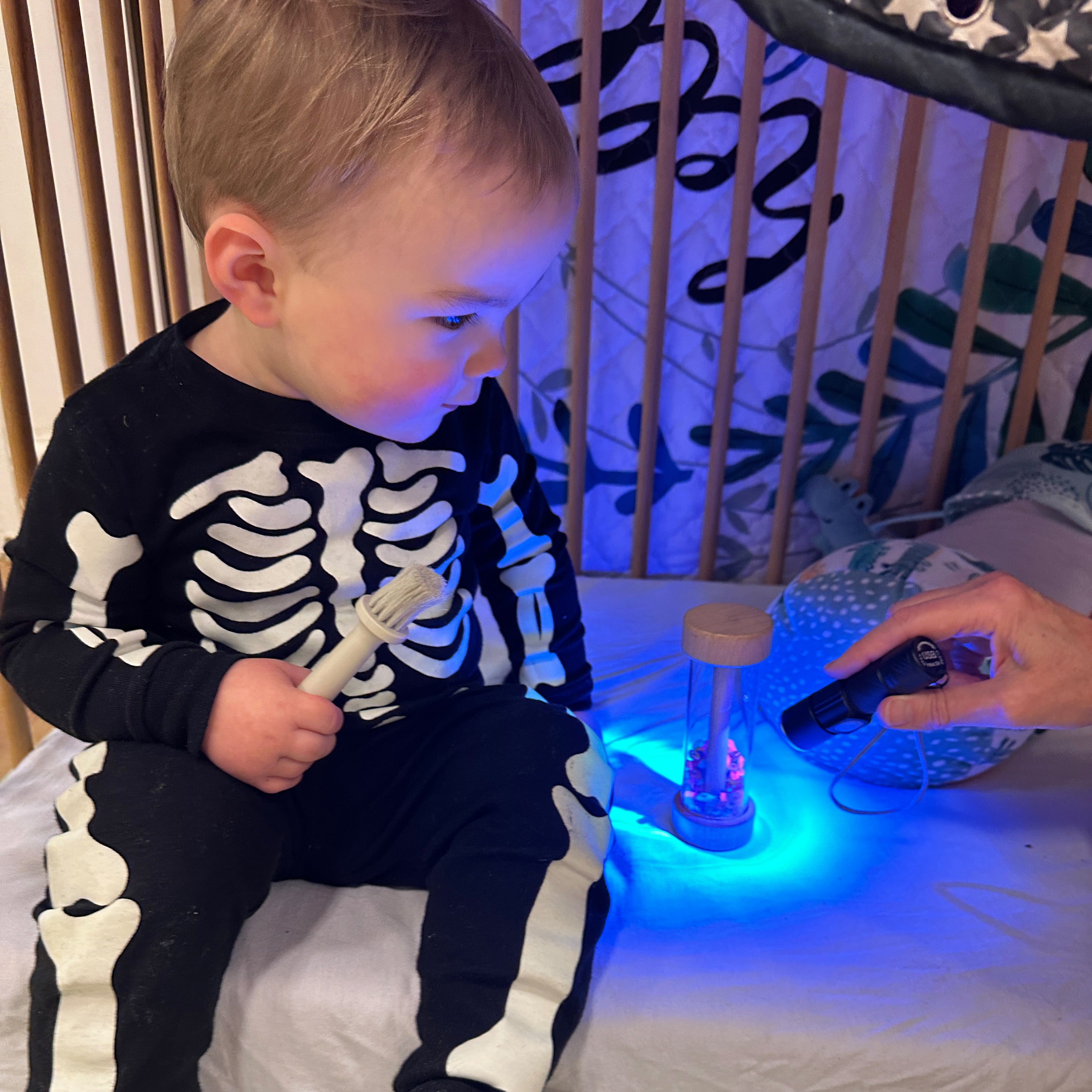 Child in skeleton pajamas playing with a glowing toy in a crib.