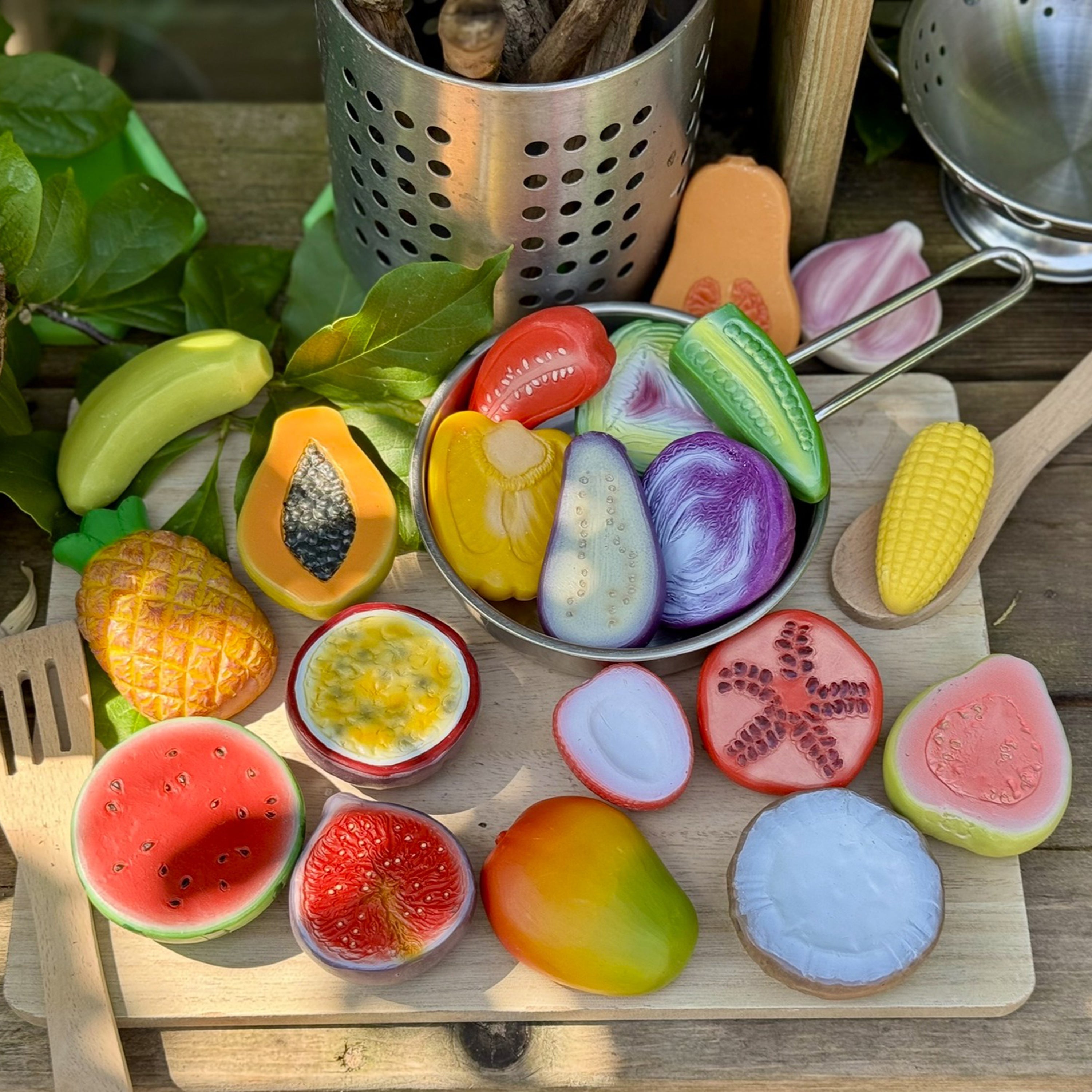 Colorful toy fruits and vegetables on a wooden surface with kitchen utensils.