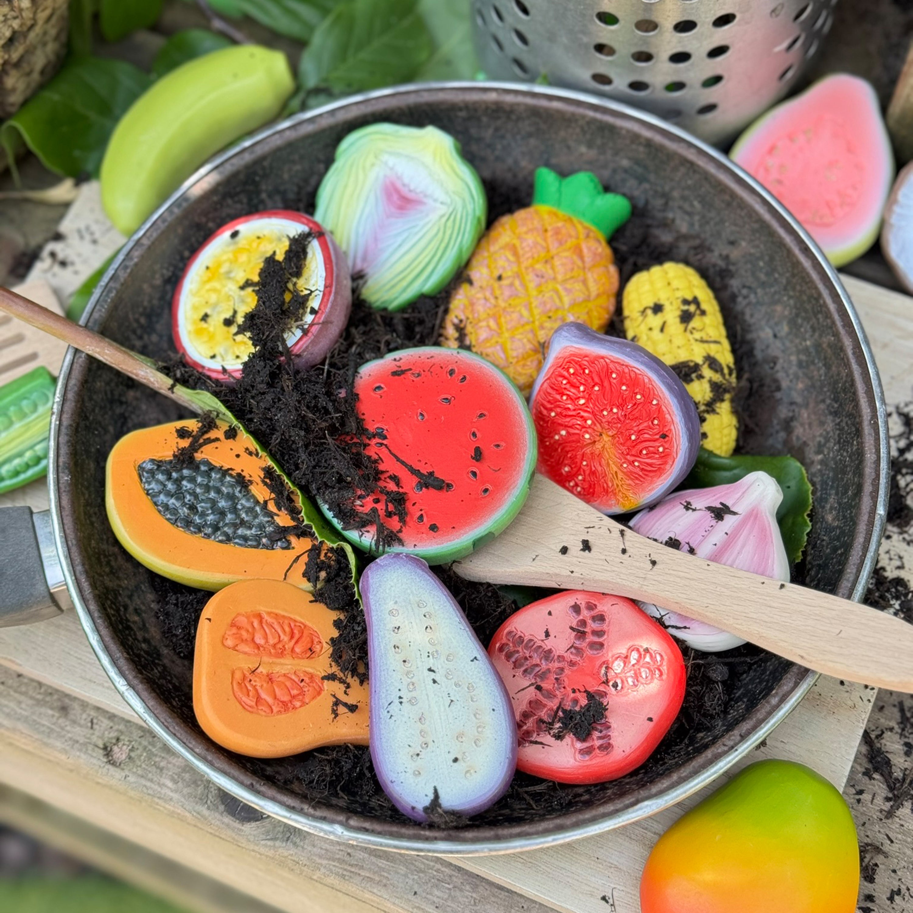 Colorful toy fruits and vegetables in a pan with wooden utensils on a rustic background